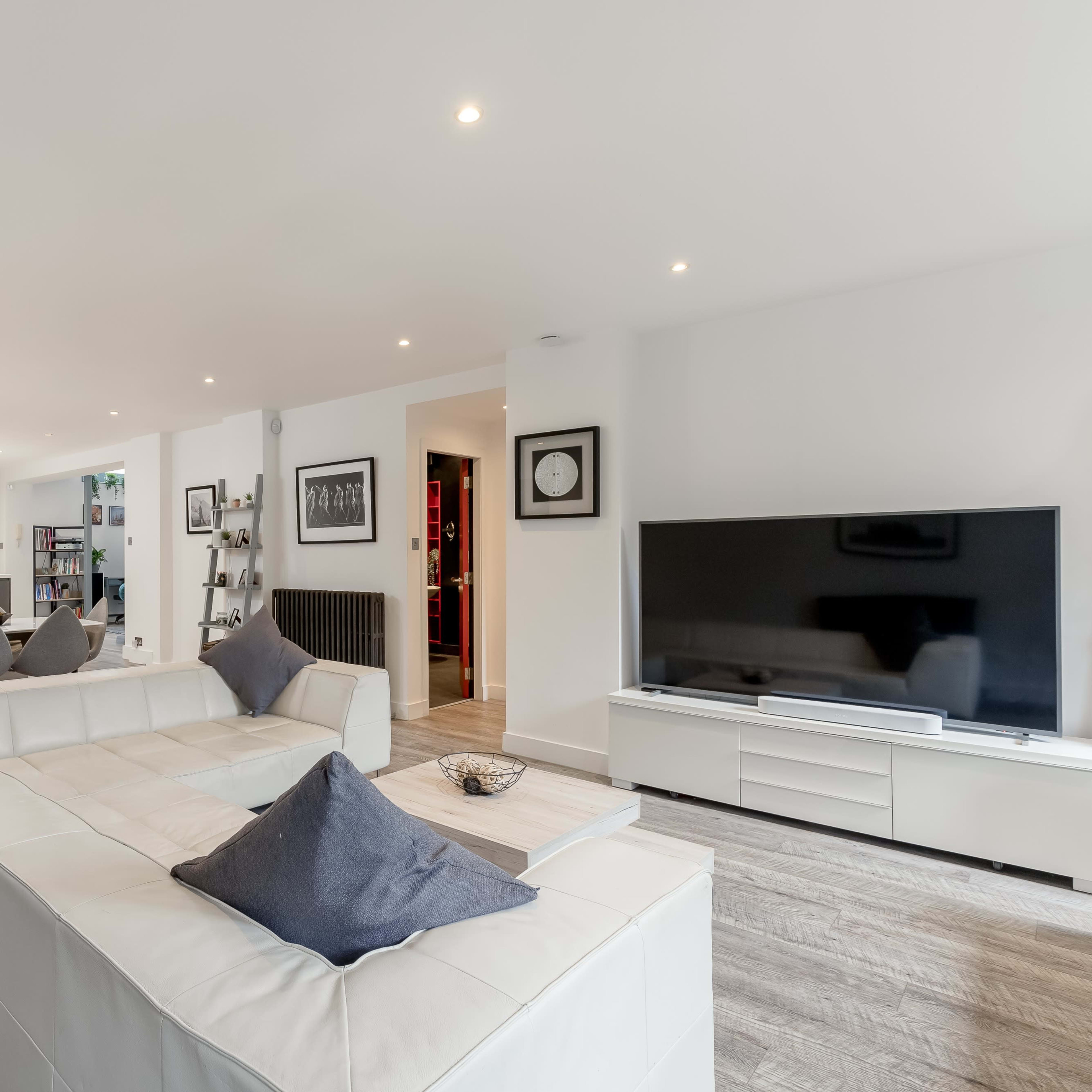 A modern living room with a white sectional sofa, a television on a white media unit, and an open layout connecting to a kitchen in the background.