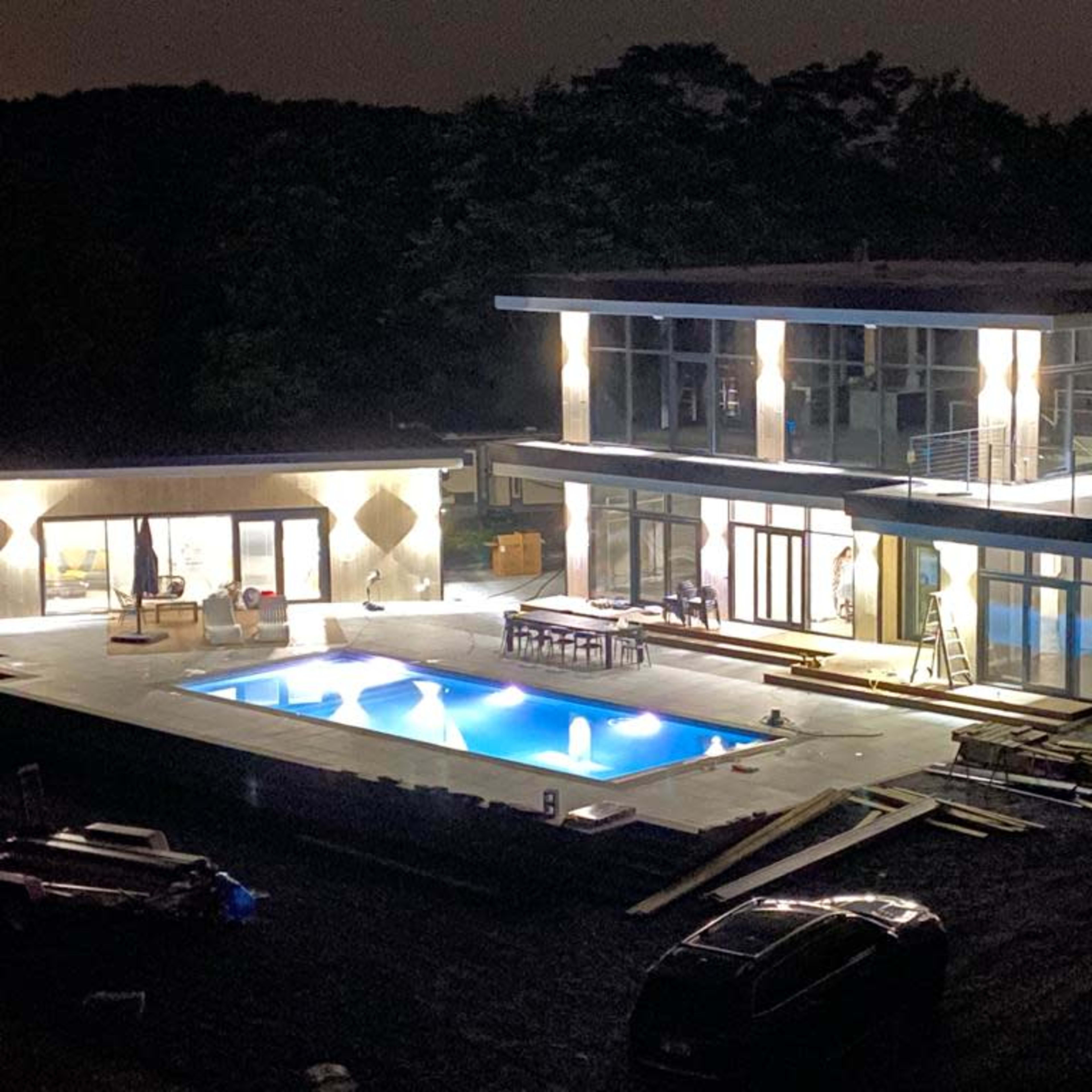 A modern two-story house with large windows and a swimming pool is illuminated at night, with construction materials and workers nearby.