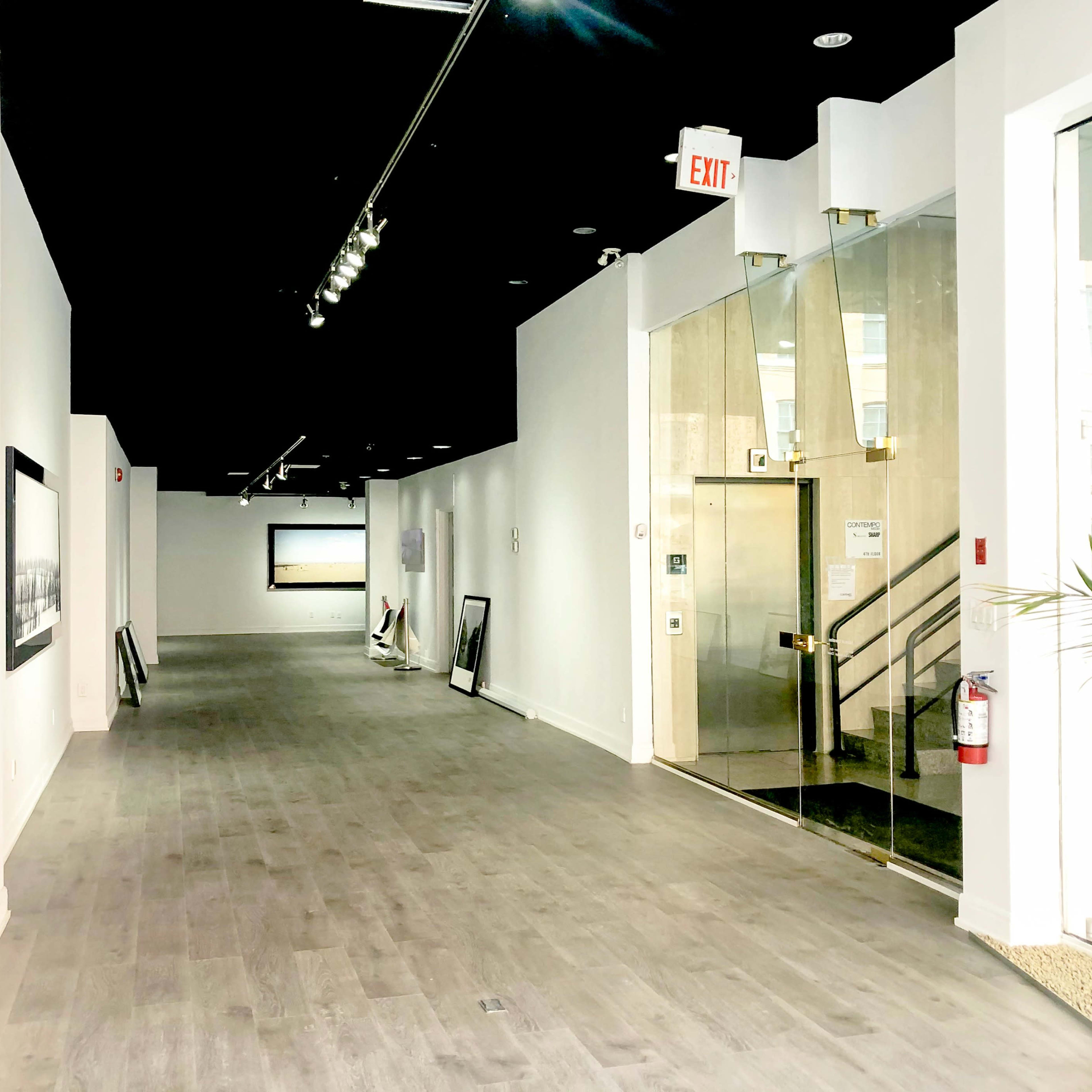 A well-lit hallway in an art gallery with large framed artworks on the walls and a glass entrance door at the end.