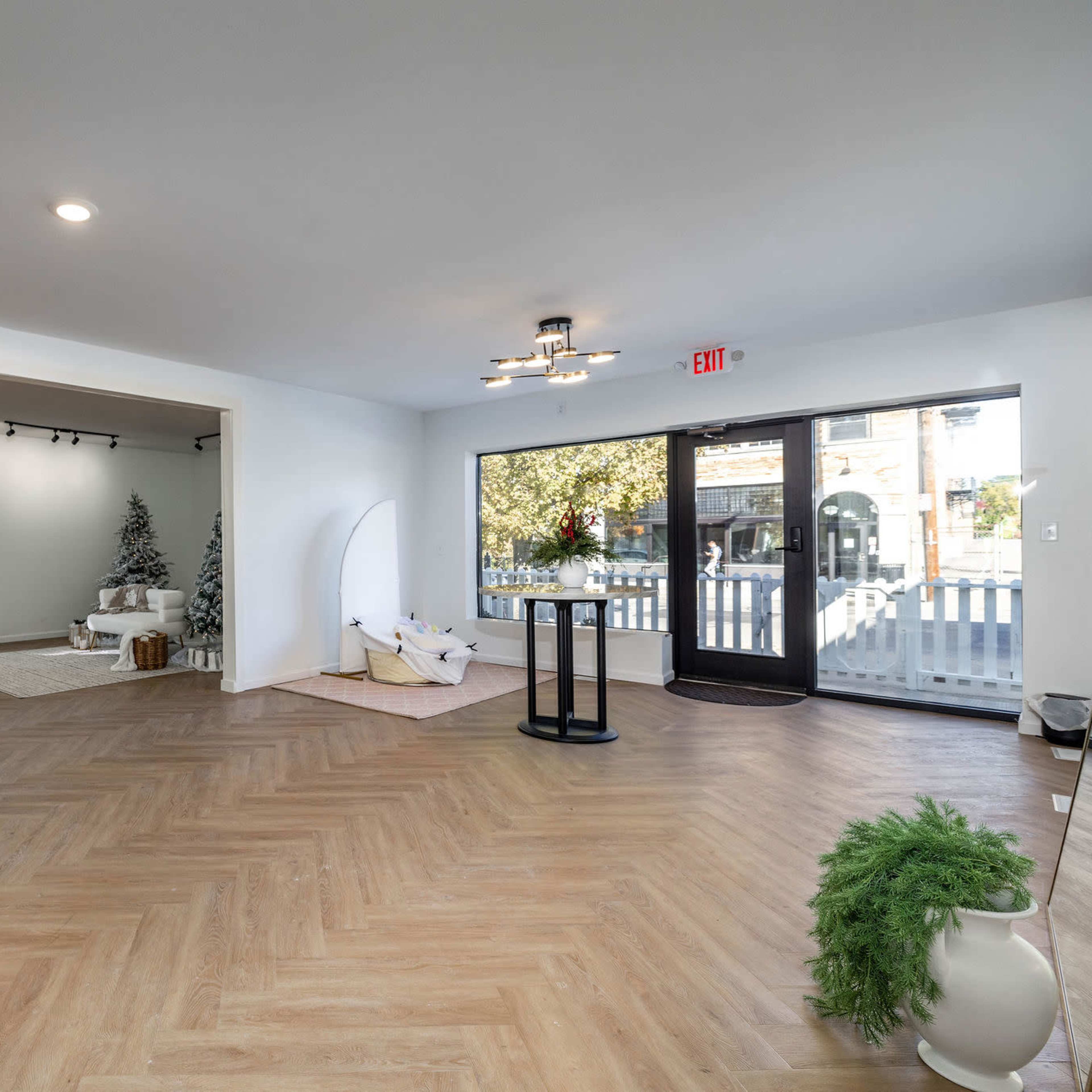 A spacious, well-lit room with wood flooring featuring a seating area, a holiday-themed corner with decorations, and large windows facing an outdoor space.