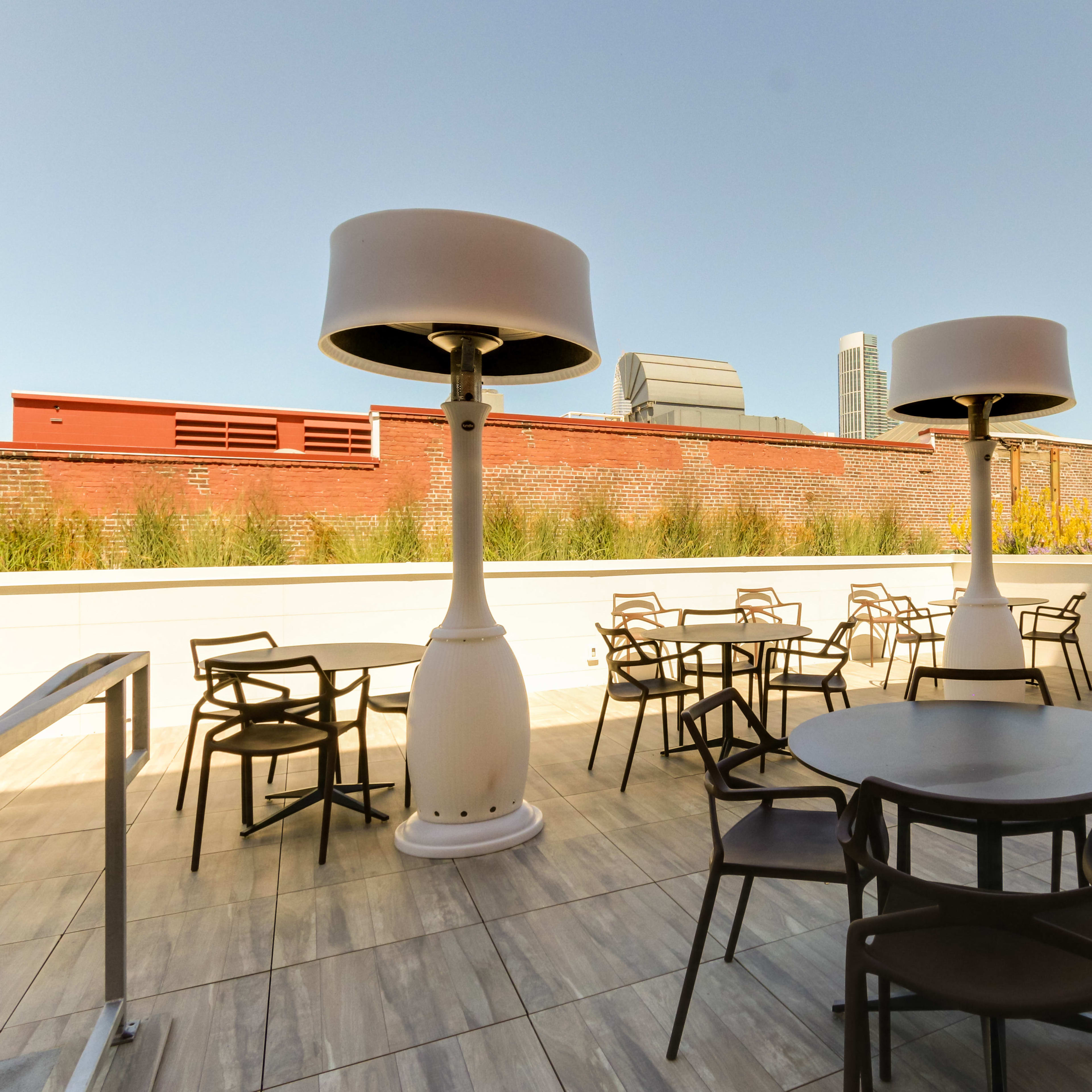 An outdoor seating area with round tables and chairs arranged around large, lamp-like structures against a backdrop of buildings and clear blue sky.