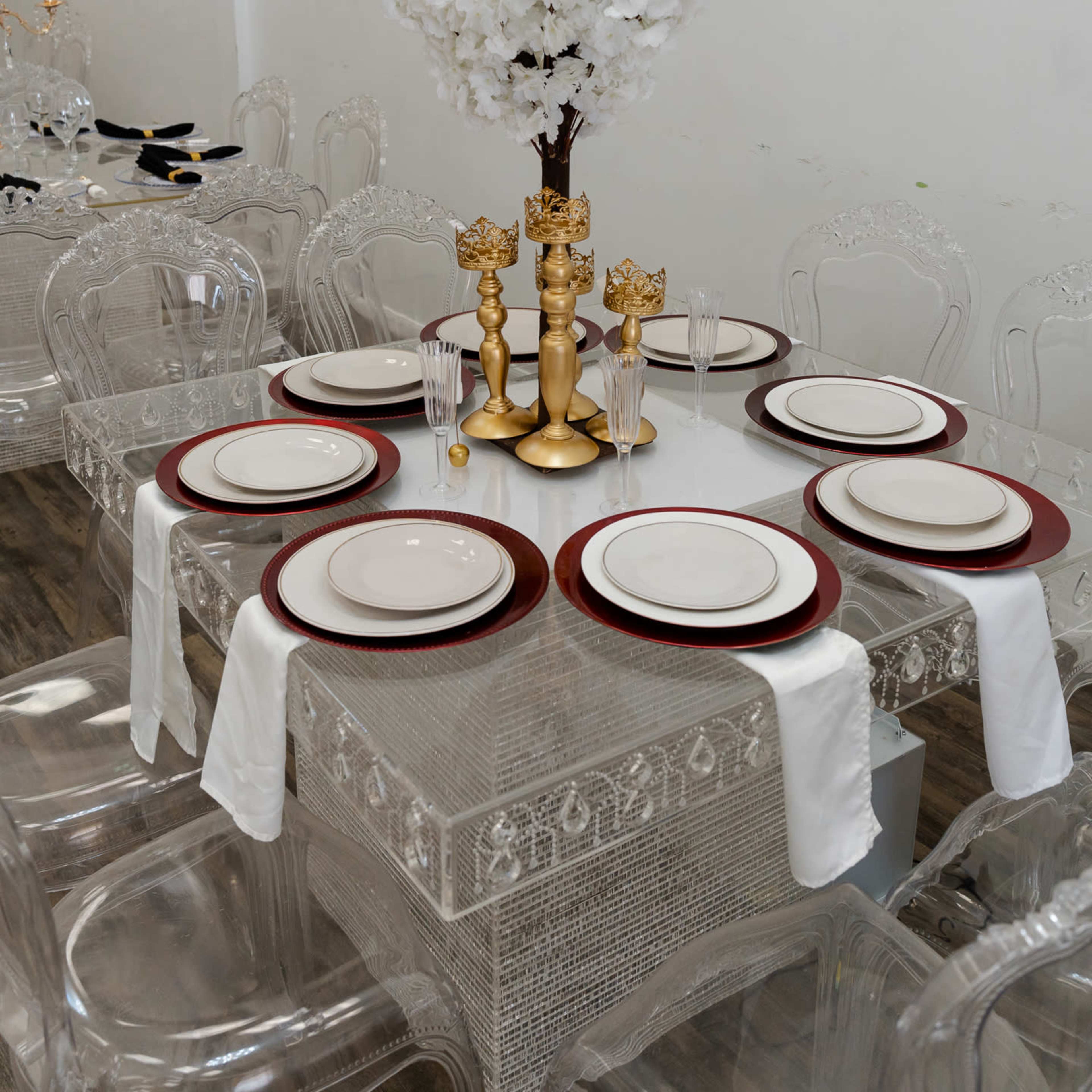A glass dining table is set with white plates on red chargers, featuring a tall arrangement of white flowers in a gold vase at the center.