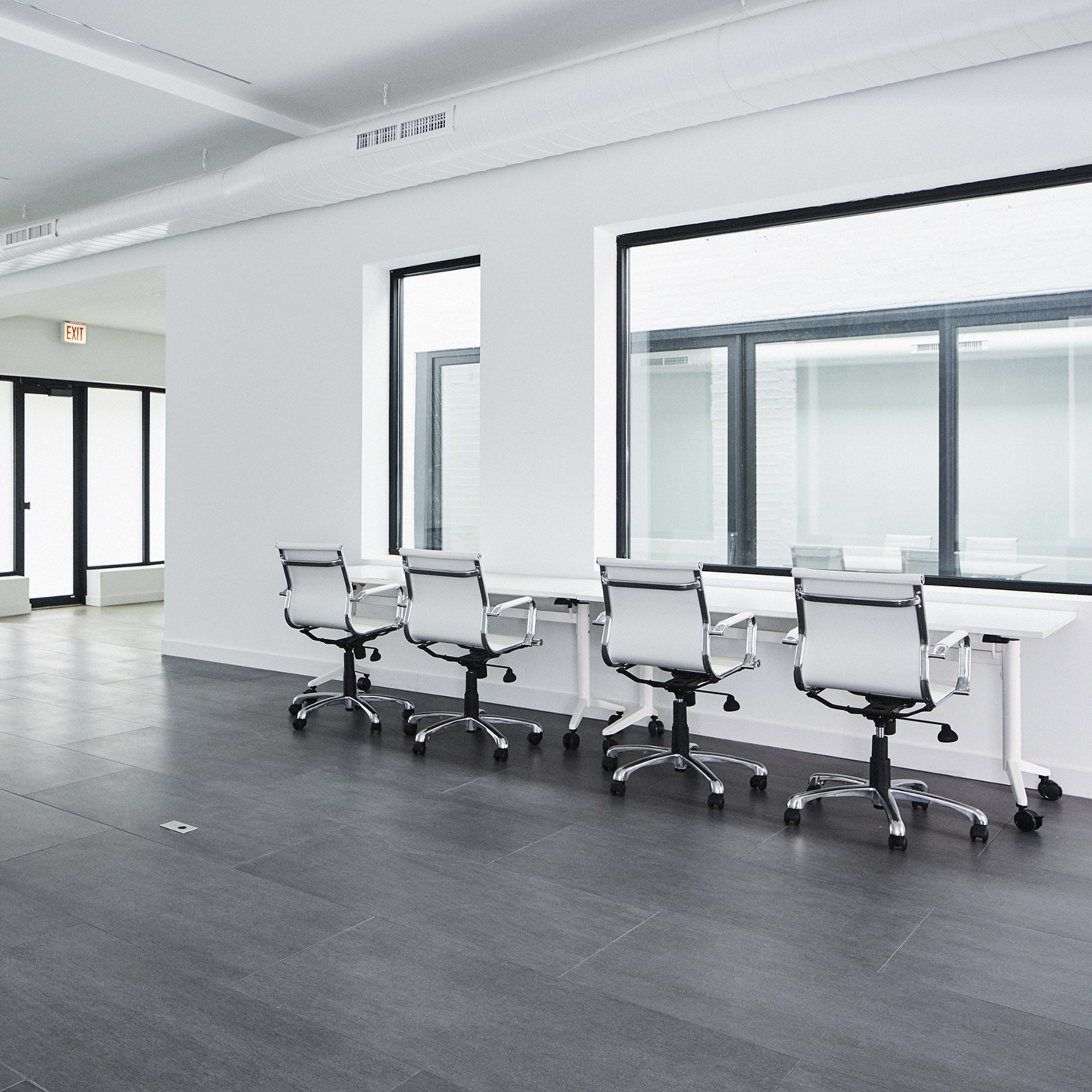 A modern, minimalist office space with a black couch and several office chairs arranged around a long desk, featuring large windows that allow natural light to enter.