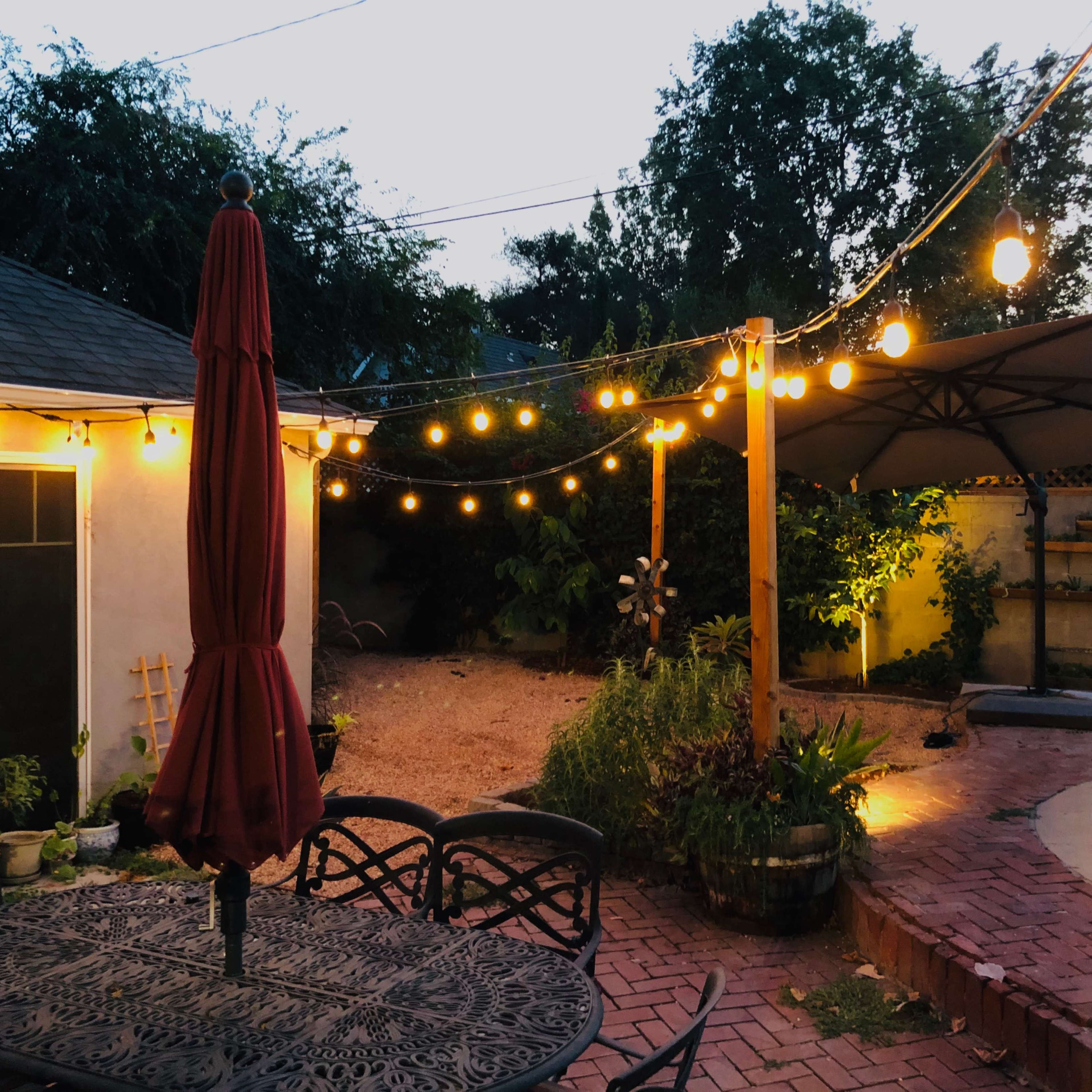 A backyard scene at dusk, featuring a patio table with a red umbrella, string lights overhead, a pool area, and greenery surrounding a small garden.