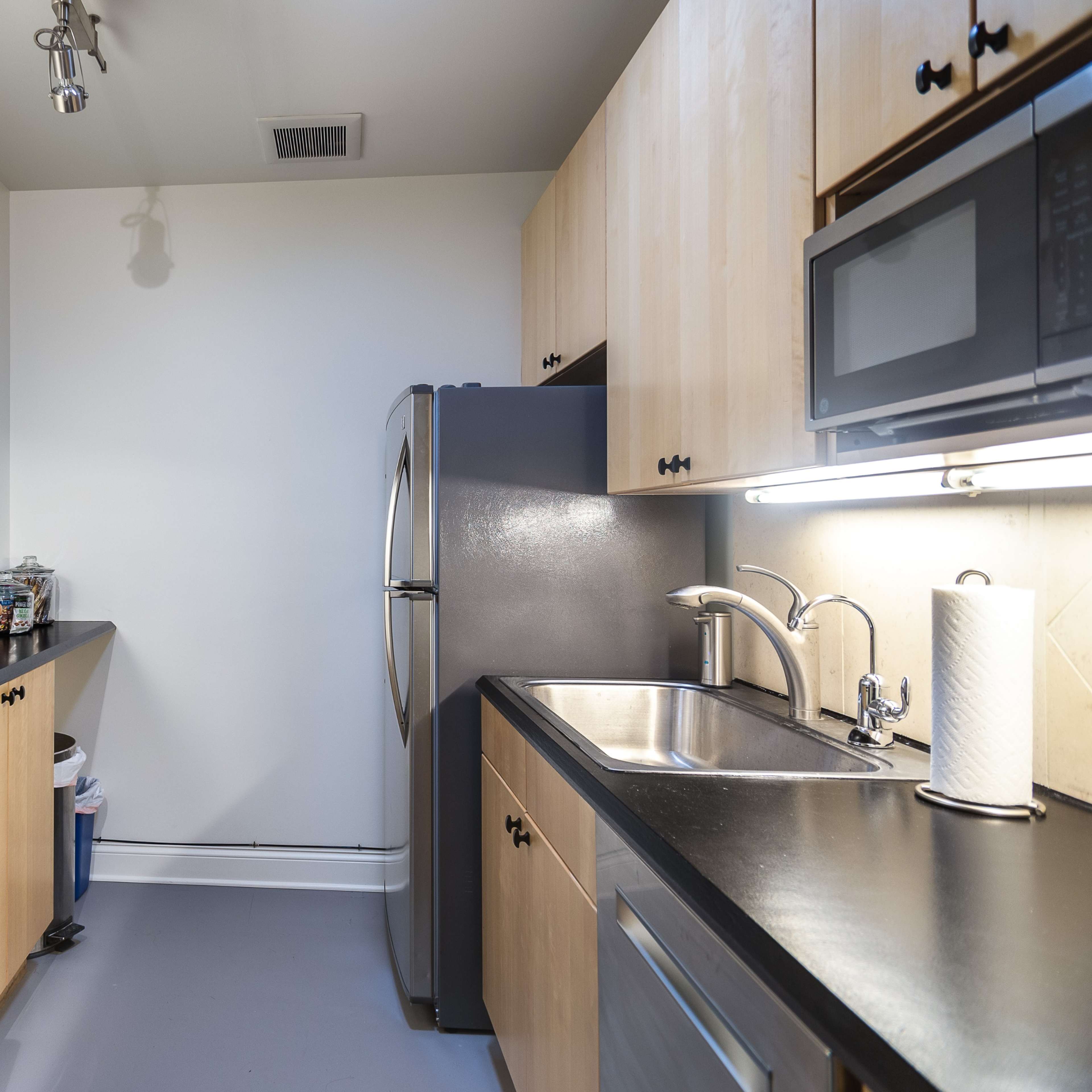 A small kitchen features black countertops, wooden cabinetry, a stainless steel refrigerator, a microwave, and an under-counter fridge.