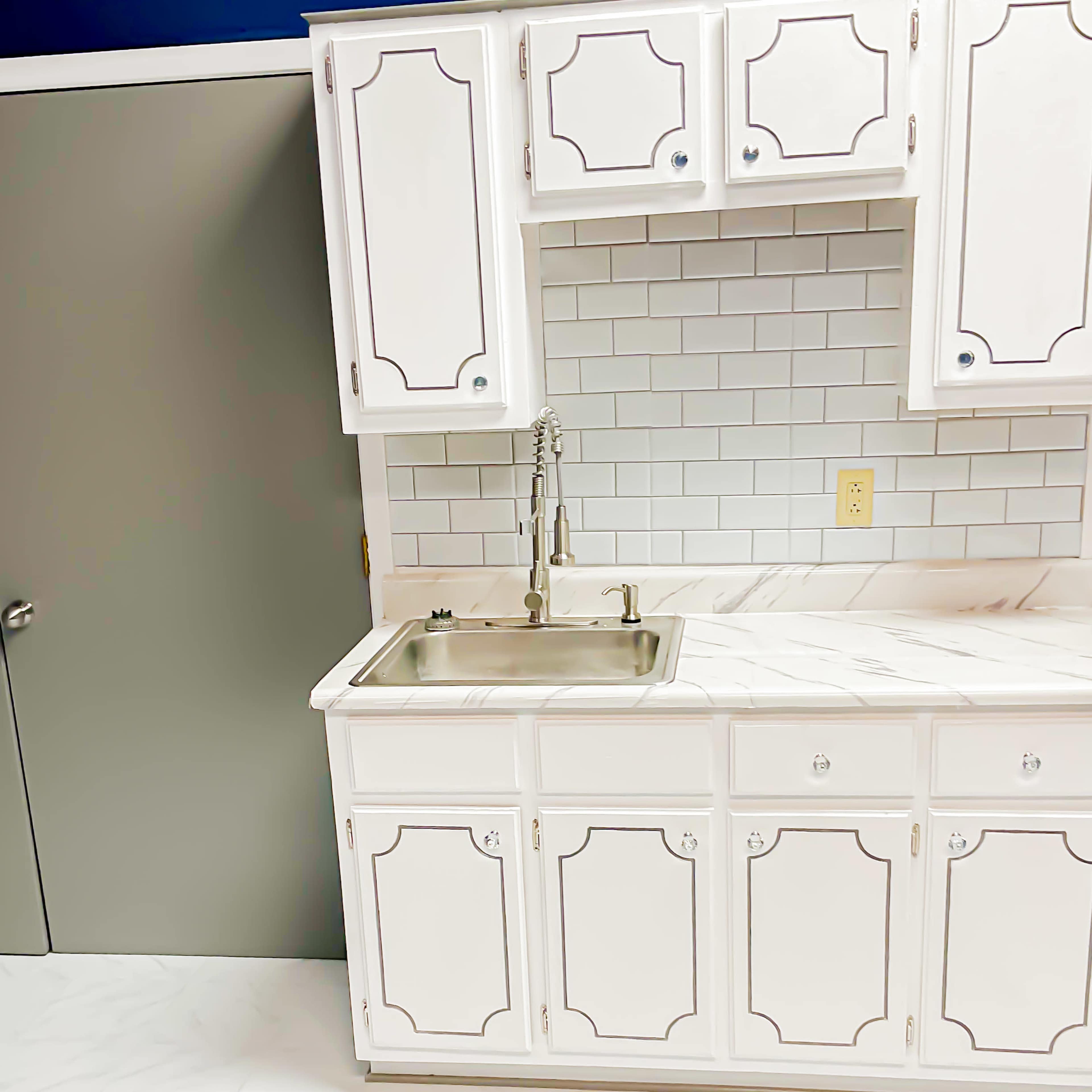 A kitchen corner with white cabinets, a stainless steel sink, and a marble-patterned countertop against a tiled backsplash.