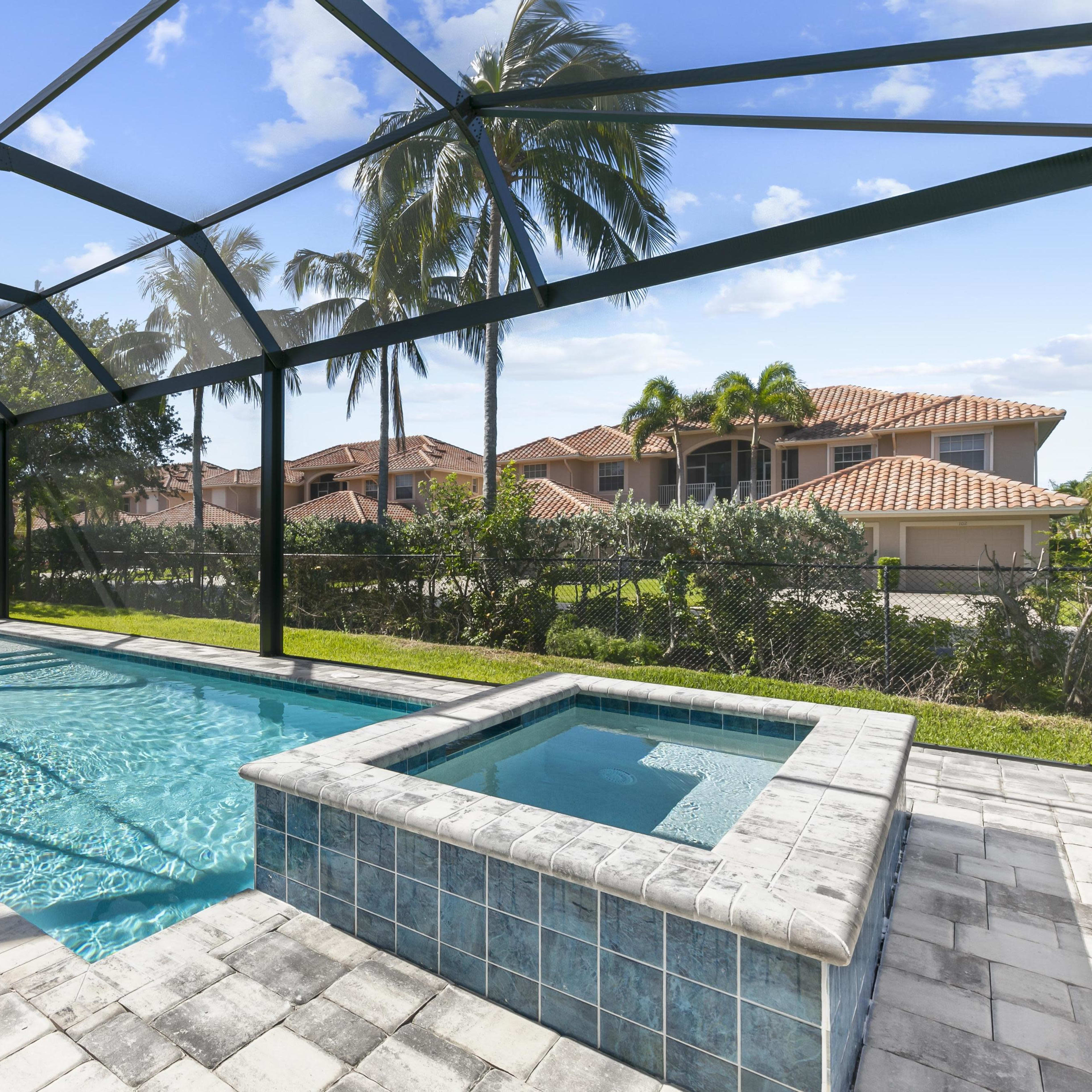 A residential backyard with a swimming pool and a hot tub, enclosed by a glass screen, surrounded by palm trees and houses in the background.