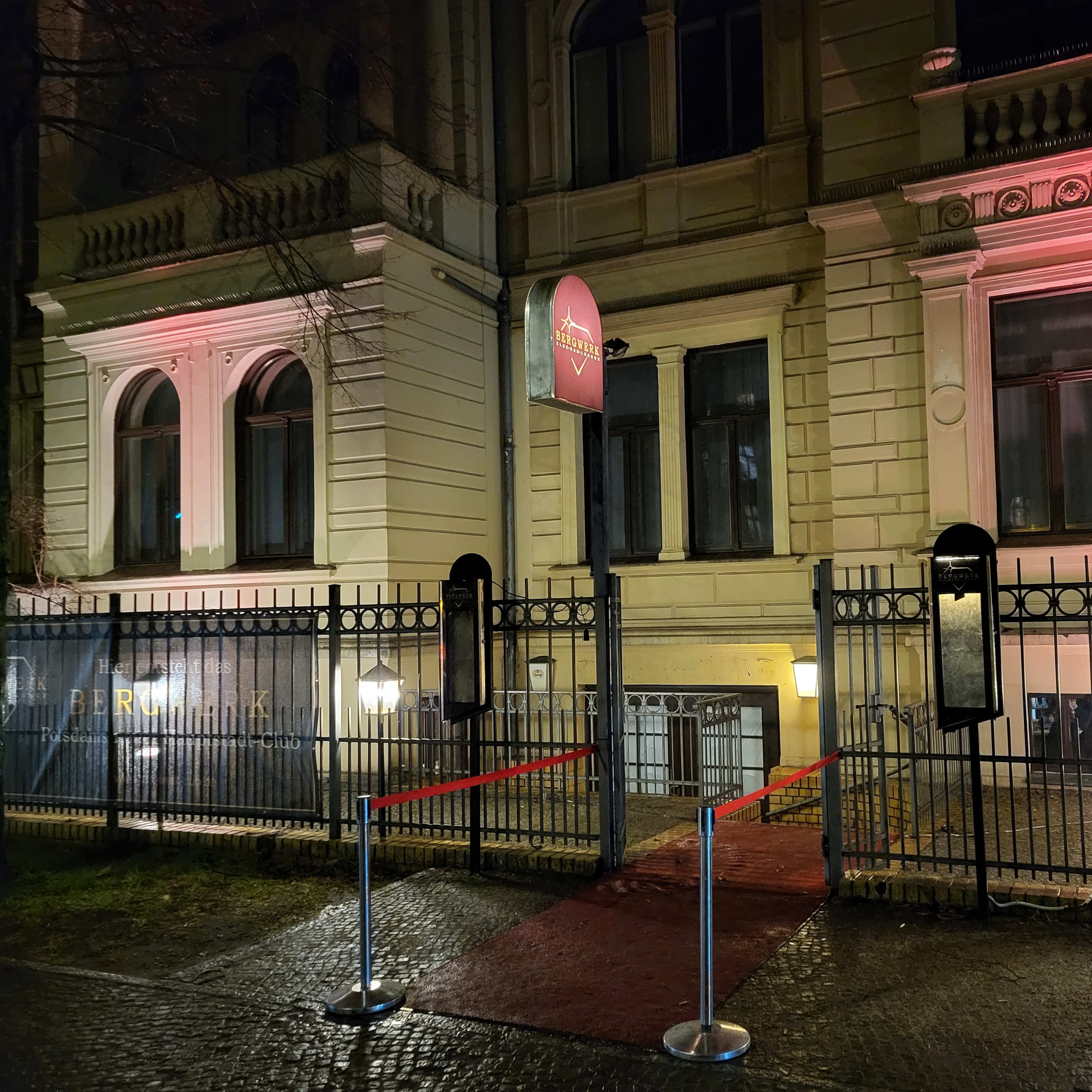The entrance of a building is framed by a gated area with red ropes and illuminated lanterns, featuring a sign that reads "Bergmark."