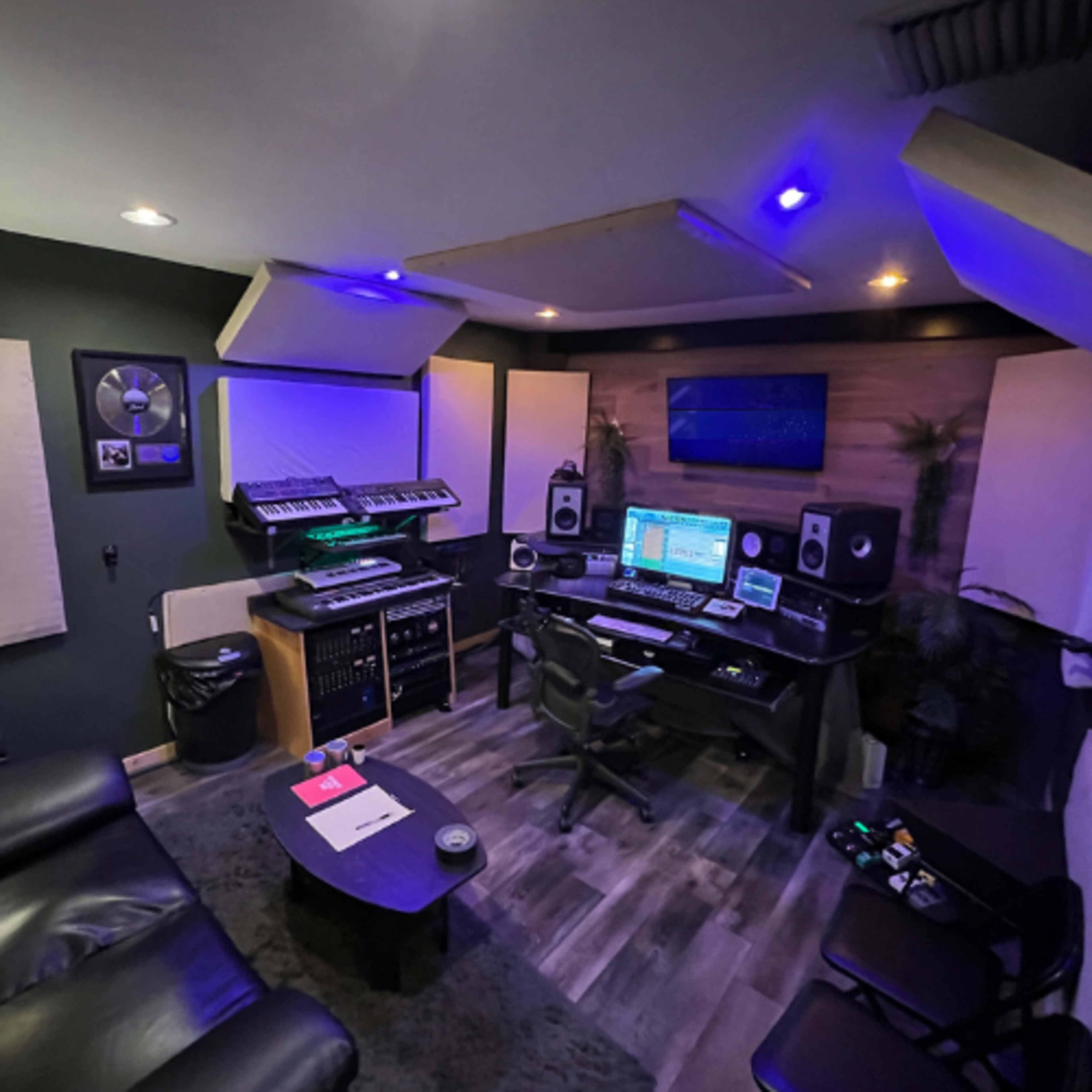 A small music production studio featuring a desk with computer equipment, synthesizers, monitors, and acoustic panels on the walls.