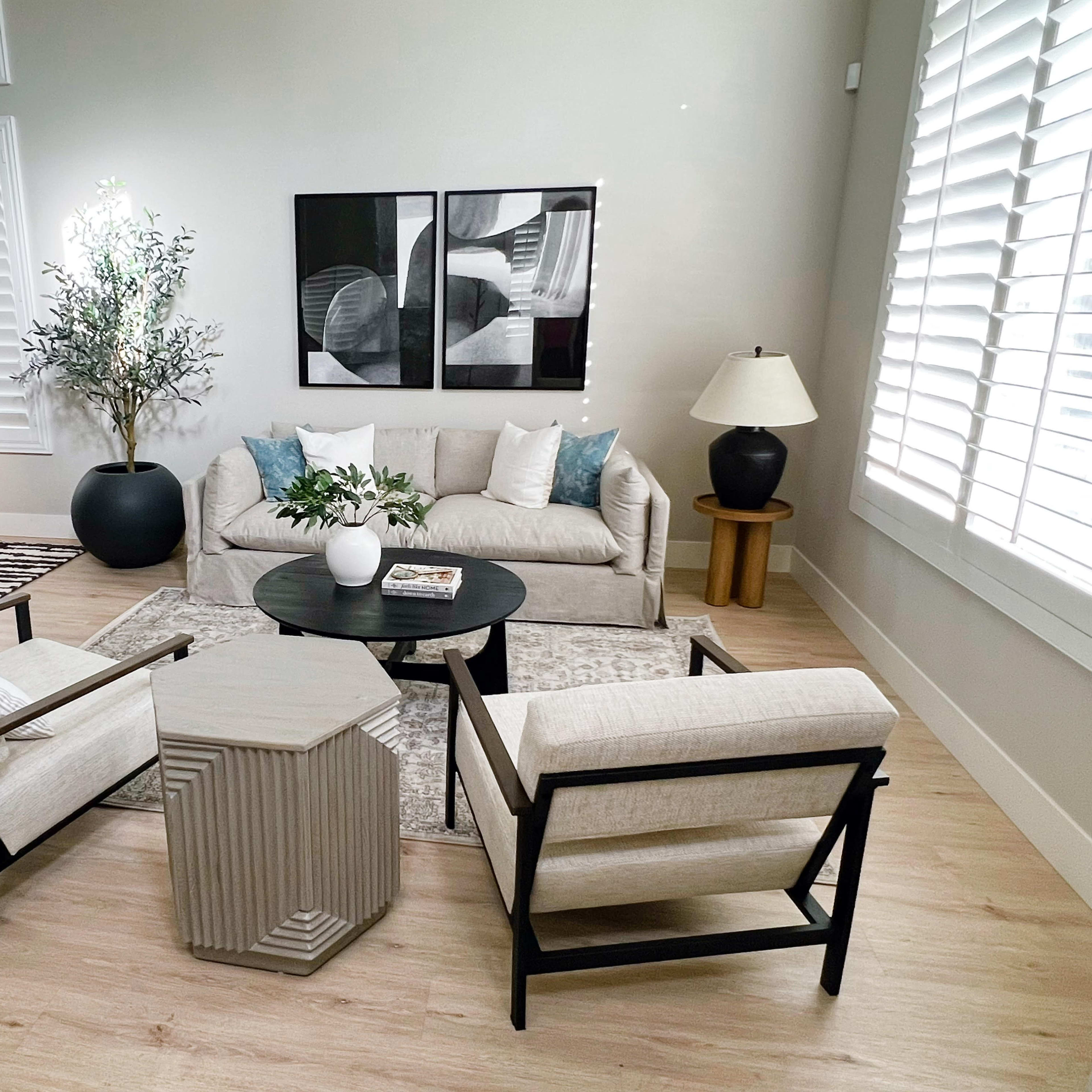 A modern living room with a light-colored sofa, two armchairs, a round coffee table, and a side table, all arranged around a rug and accompanied by decorative plants and artwork on the walls.