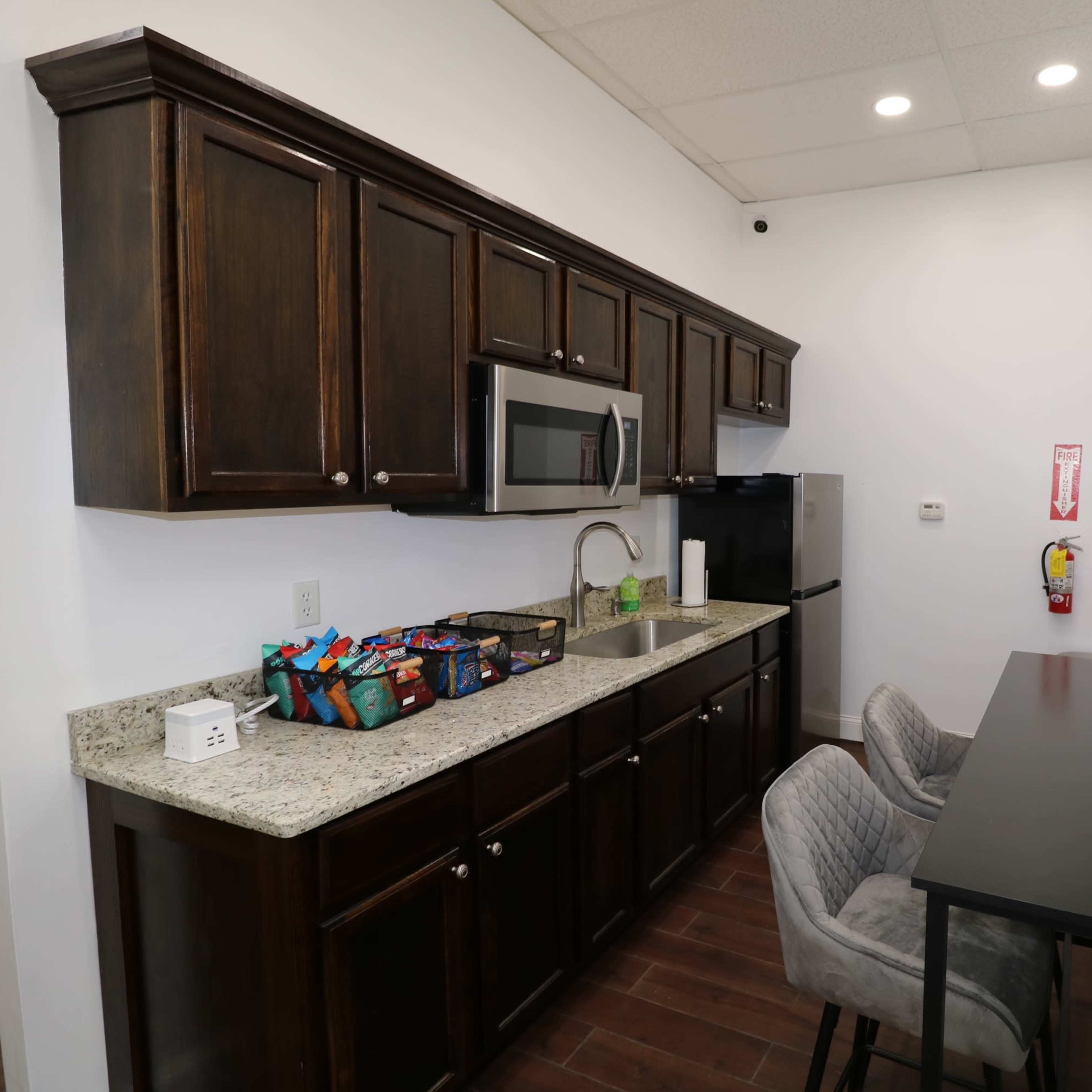 A compact kitchen with dark wooden cabinets, a granite countertop, a microwave, and a small dining table with two gray chairs.