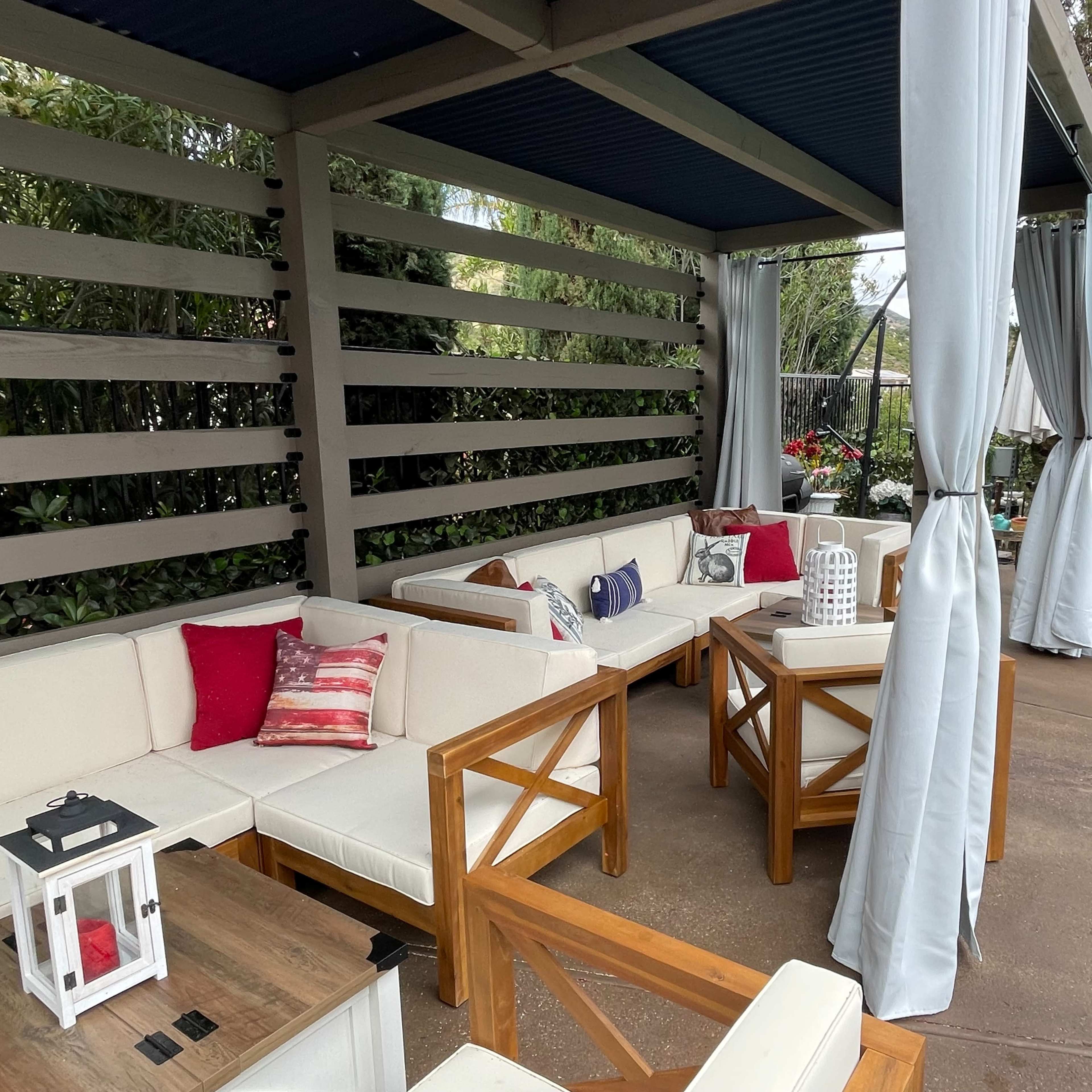 A covered outdoor seating area with white sofas and wooden tables, surrounded by greenery.