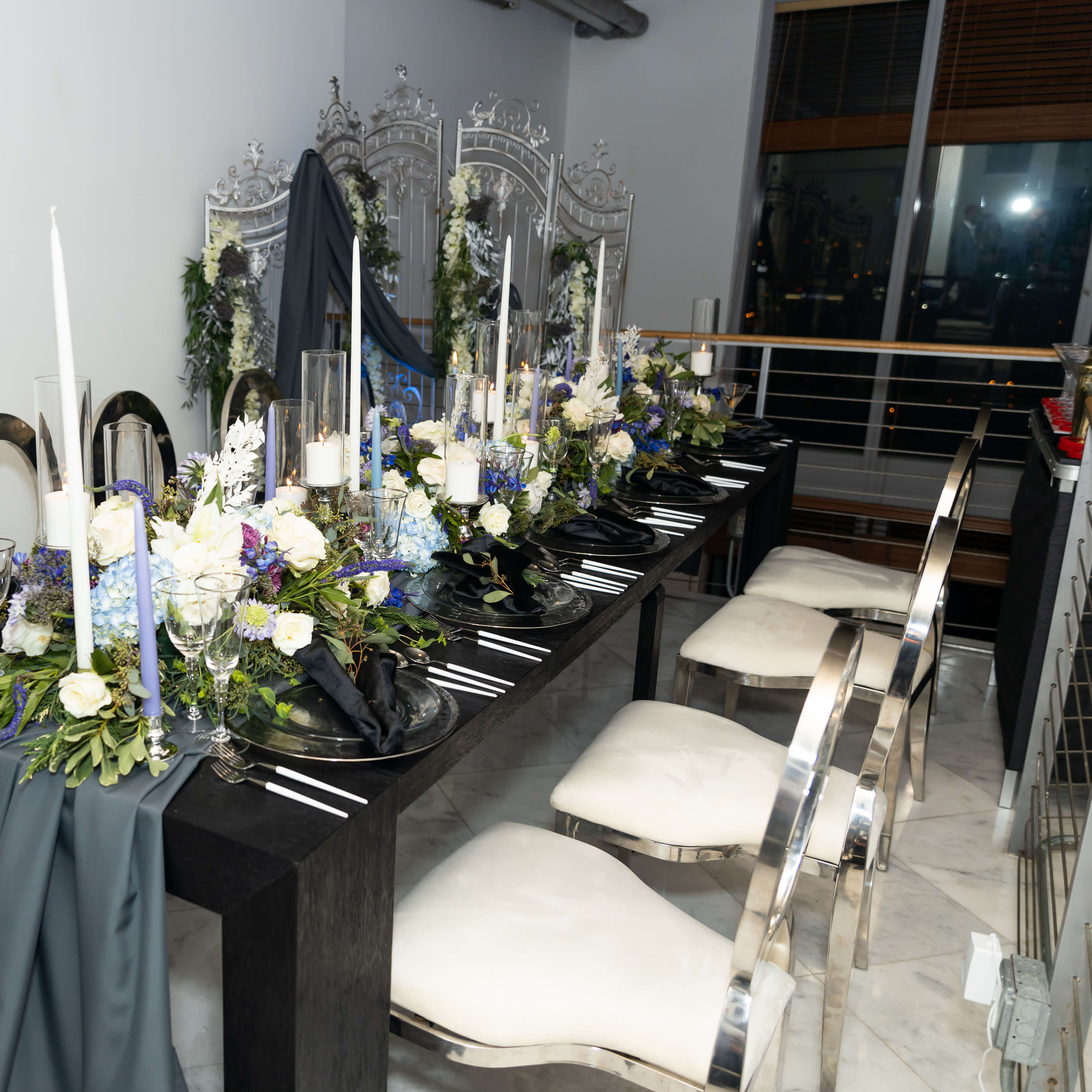 A long dining table is set with elegant floral arrangements, candles, and silverware in a modern interior space.
