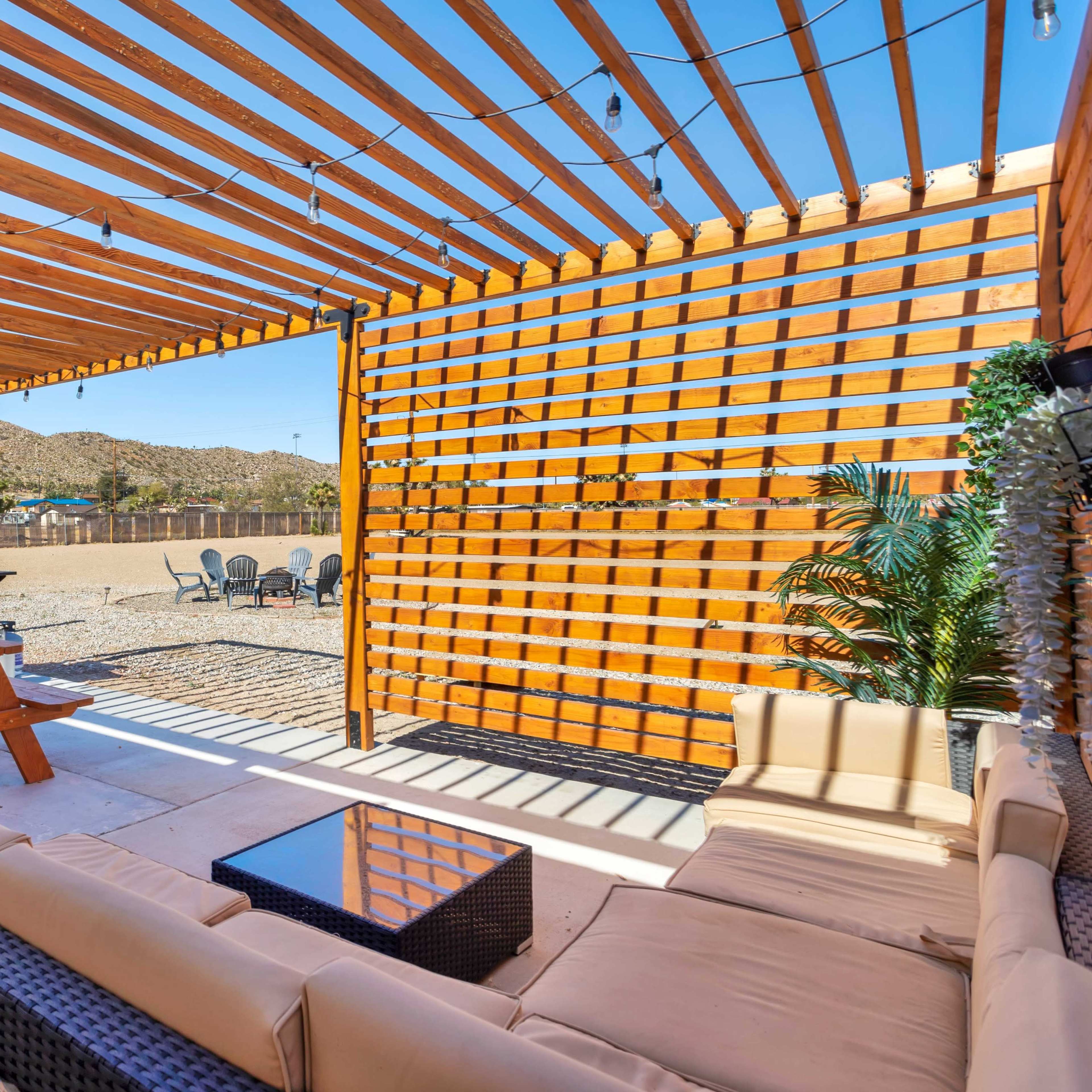 A patio area with a wooden pergola, picnic table, outdoor seating, and a view of a gravel yard with a hammock and chairs in the background.