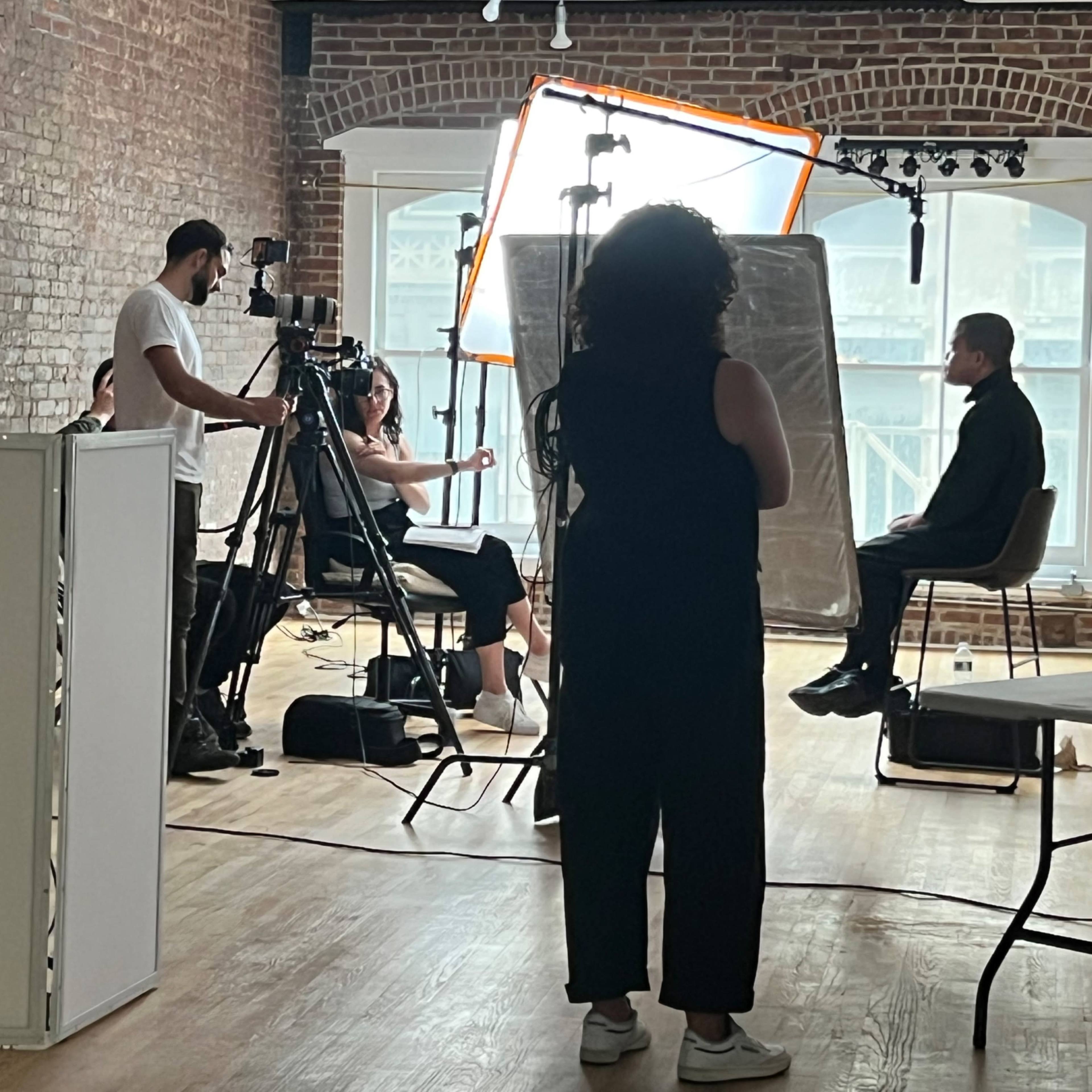 A camera crew films an interview or photoshoot in a studio with exposed brick walls and large windows.