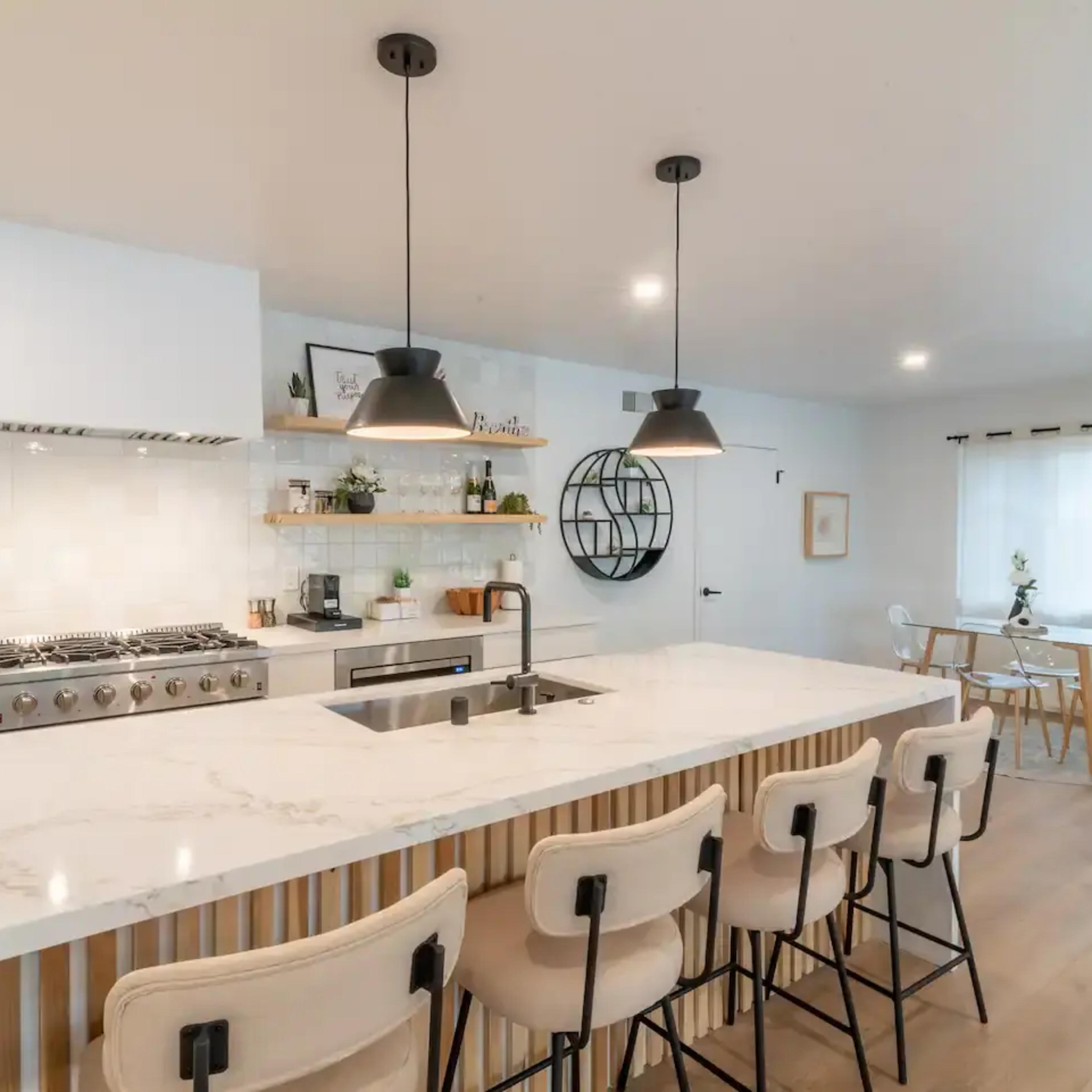 A modern kitchen with a large marble island, four bar stools, and a dining area featuring a round table and plants.
