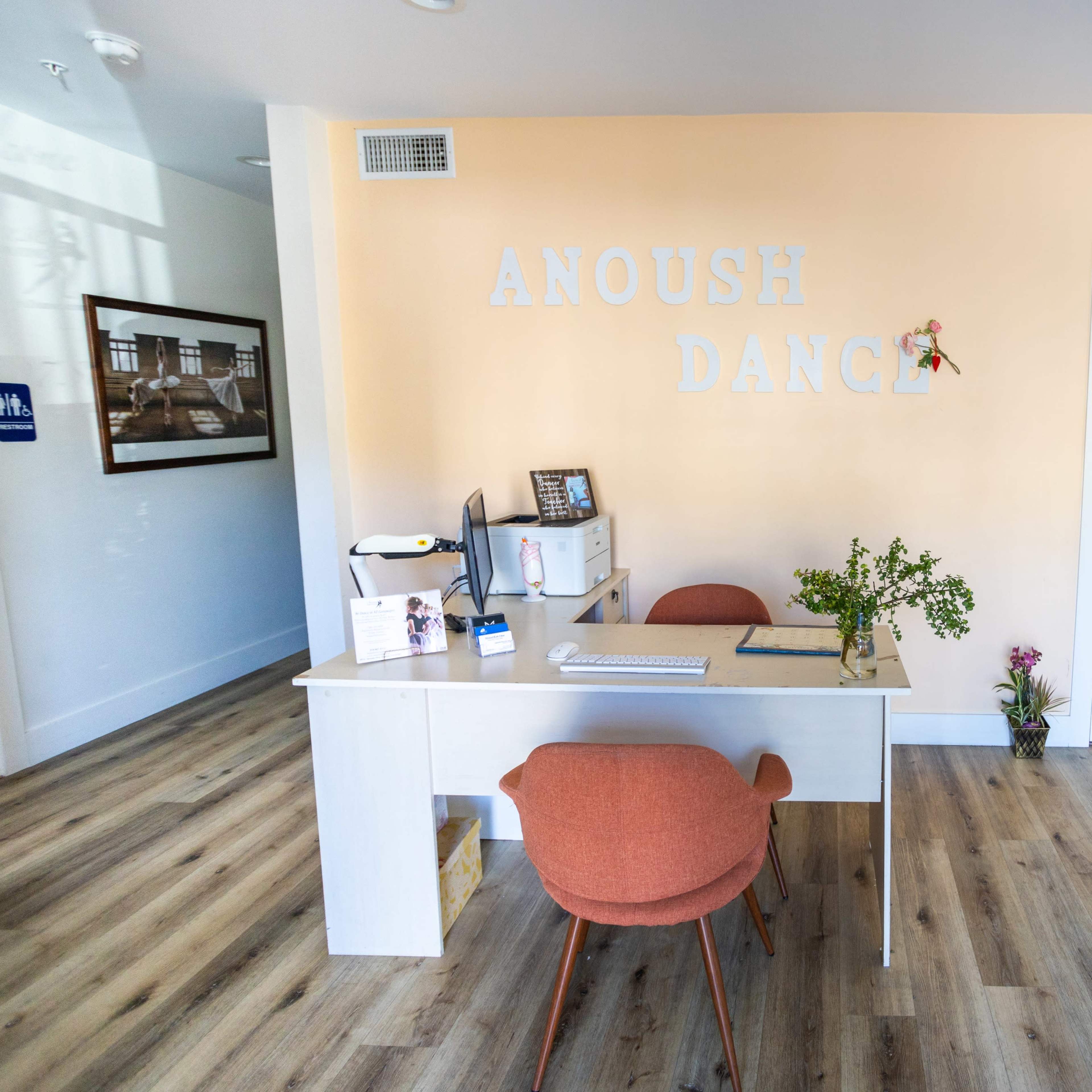 A bright and modern dance studio reception area with a white desk, two chairs, and a decorative wall featuring the words "ANOUSh DANCE."