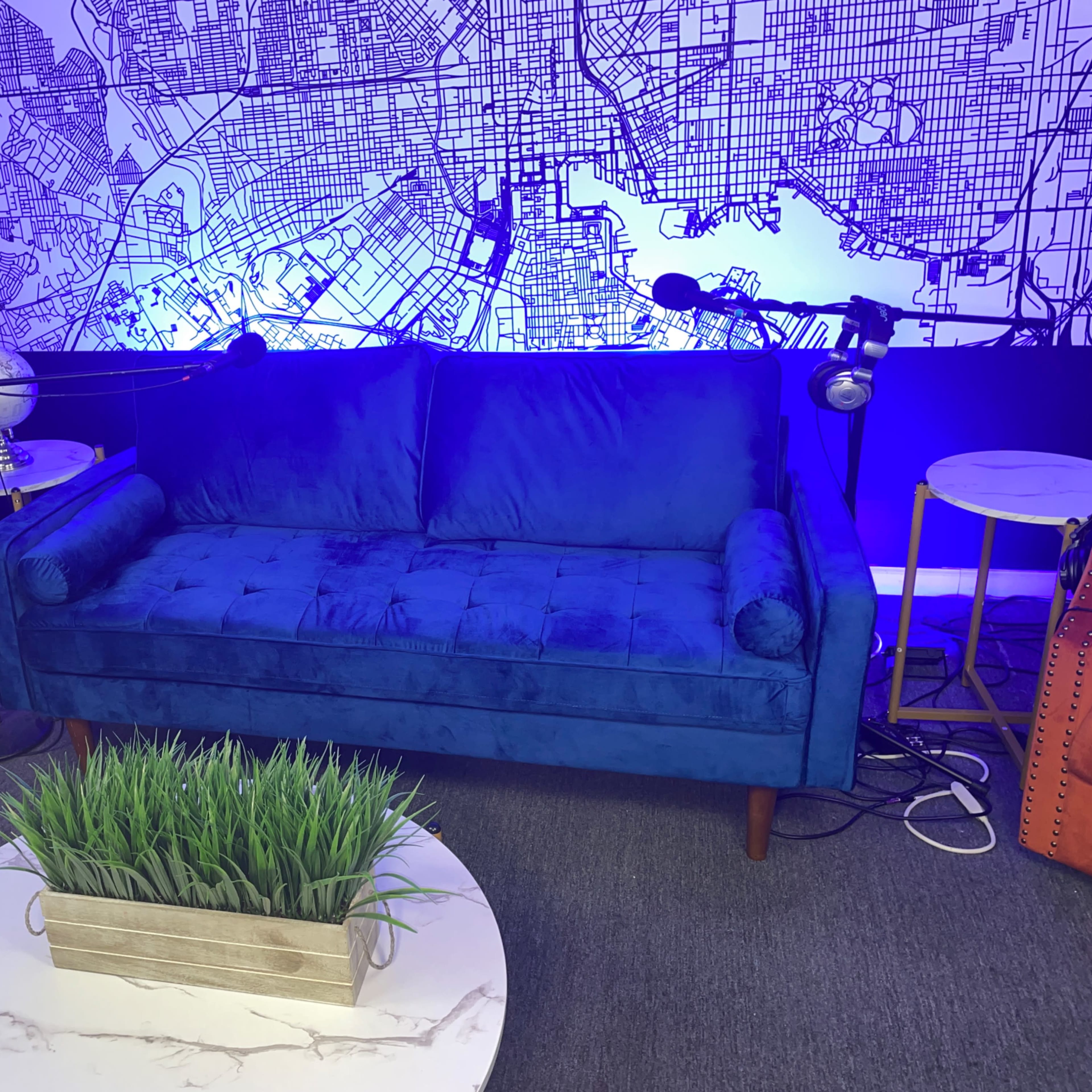 A blue couch is positioned in front of a large wall map, accompanied by two chairs, a coffee table with a planter, and several microphones on stands.