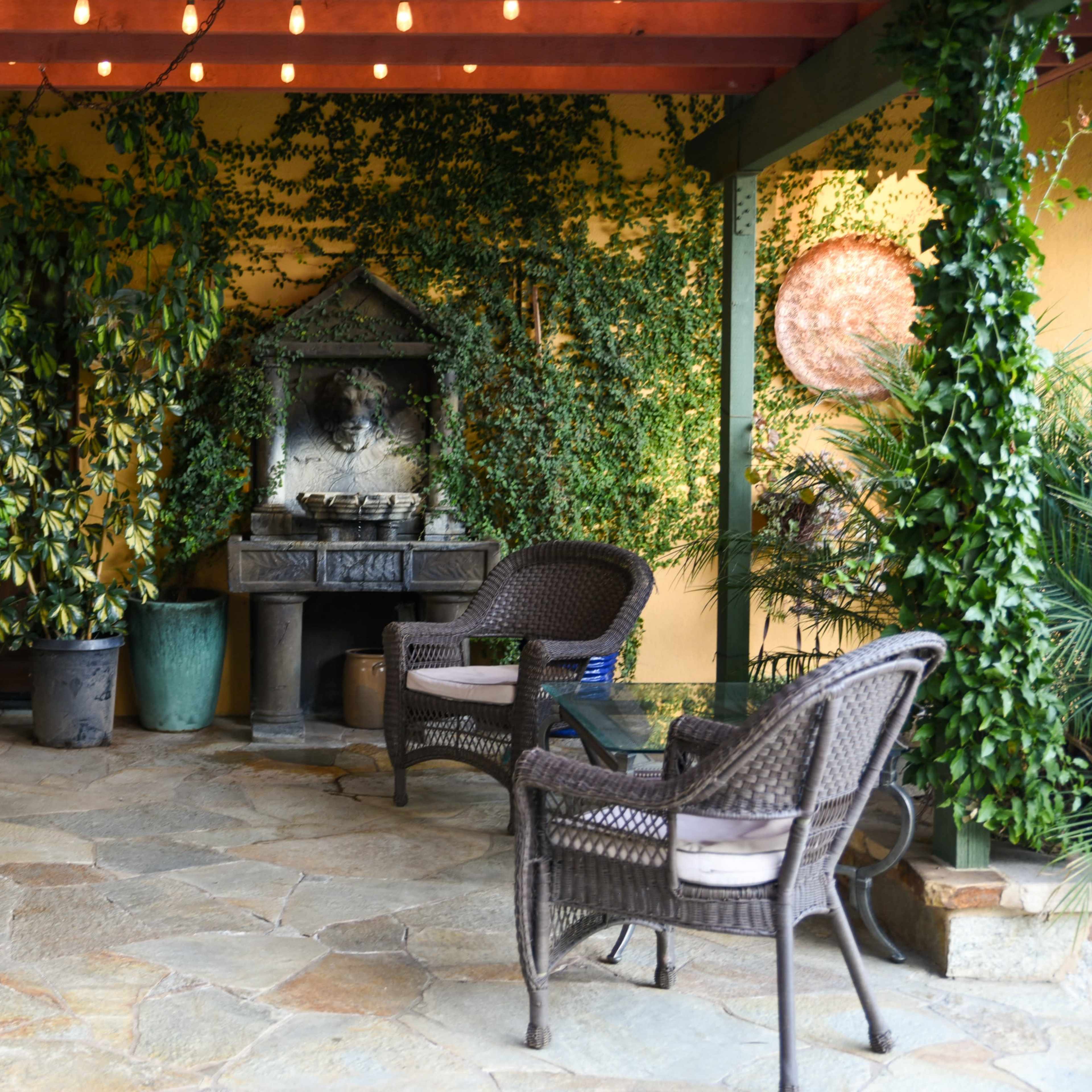 A shaded patio area with two wicker chairs, a fountain surrounded by greenery, and a stone floor.