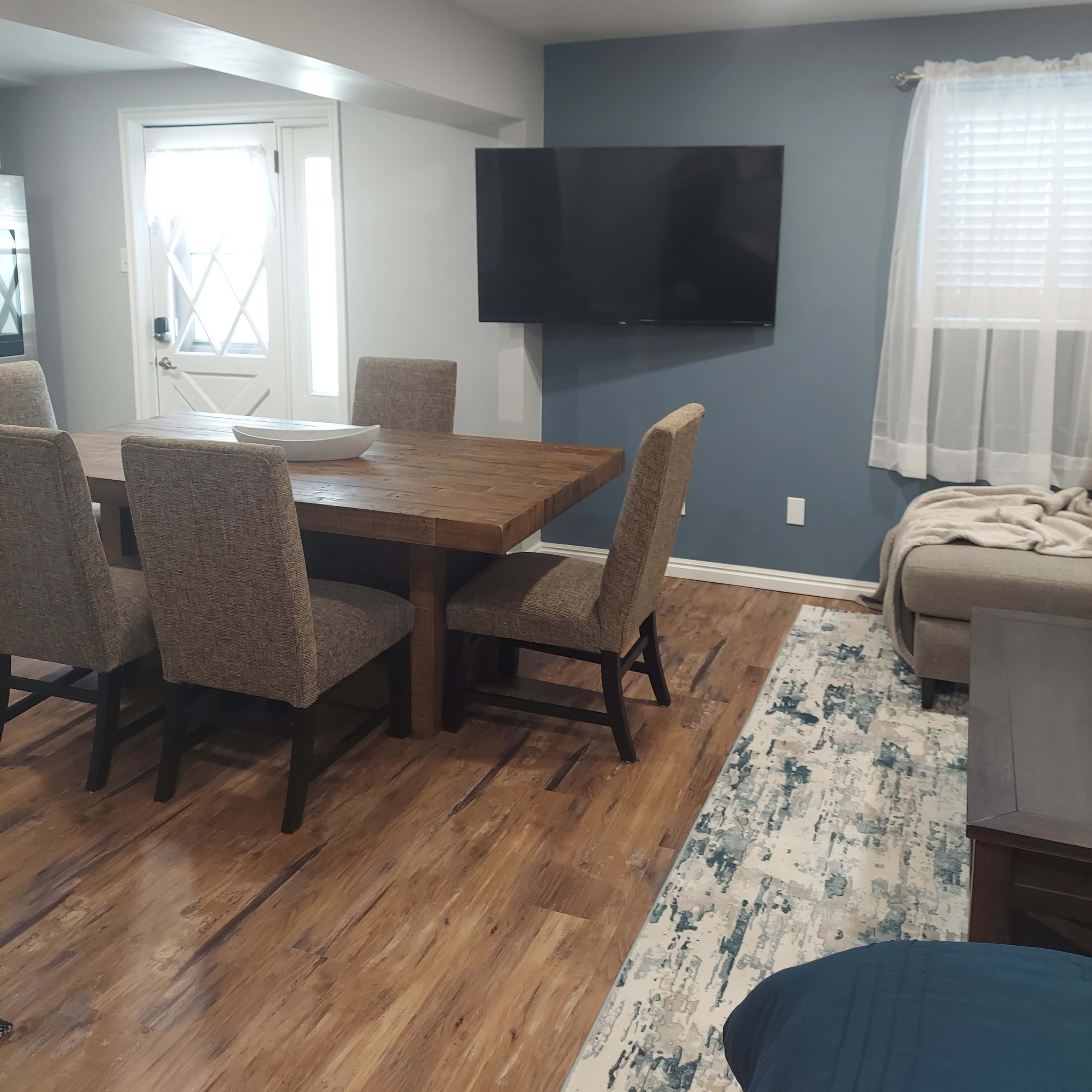 A dining area with a wooden table and six upholstered chairs, adjacent to a living space featuring a sofa, television mounted on a blue wall, and a window with sheer curtains.