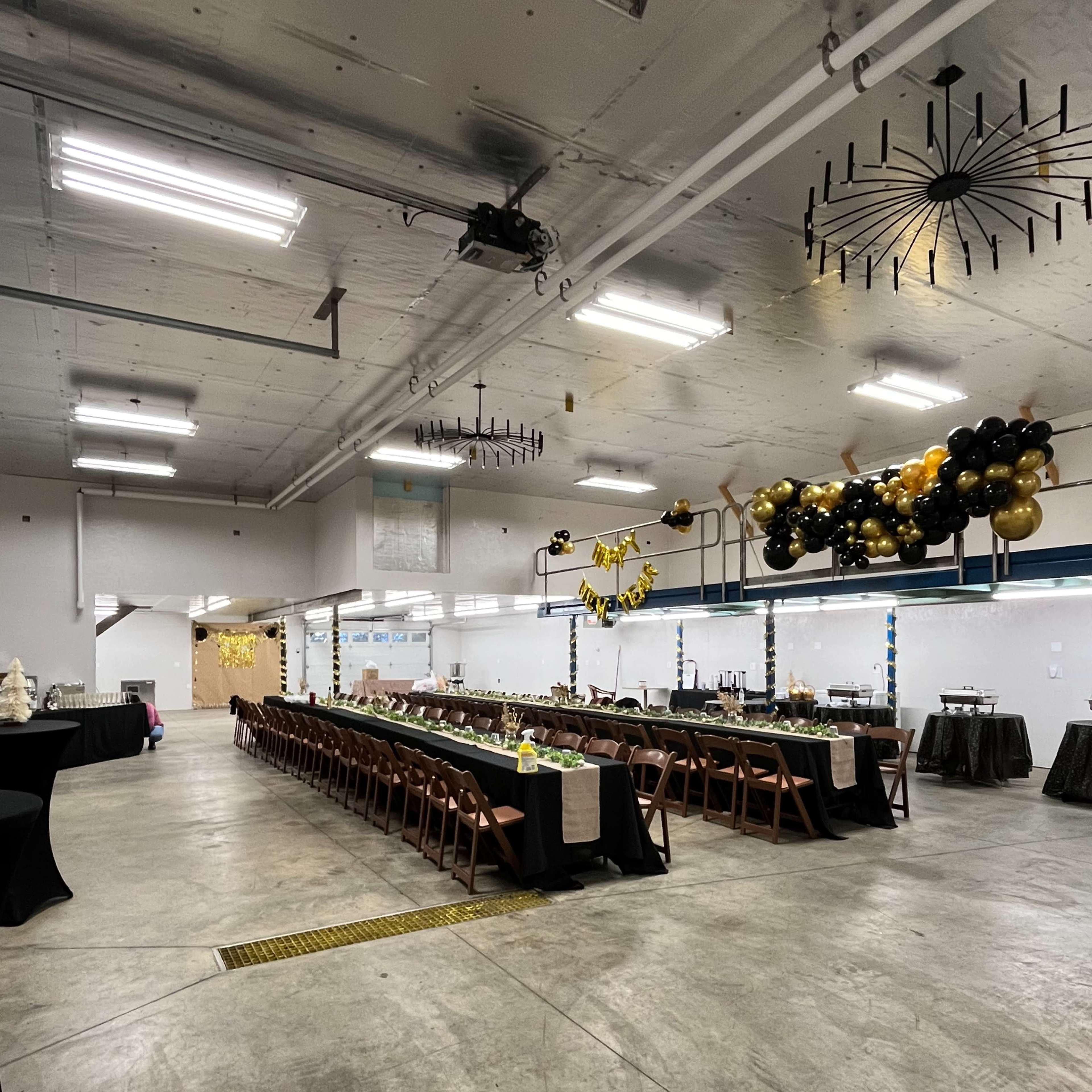 A spacious indoor venue features long tables arranged for a banquet, decorated with black and gold balloons and additional tables set for serving.