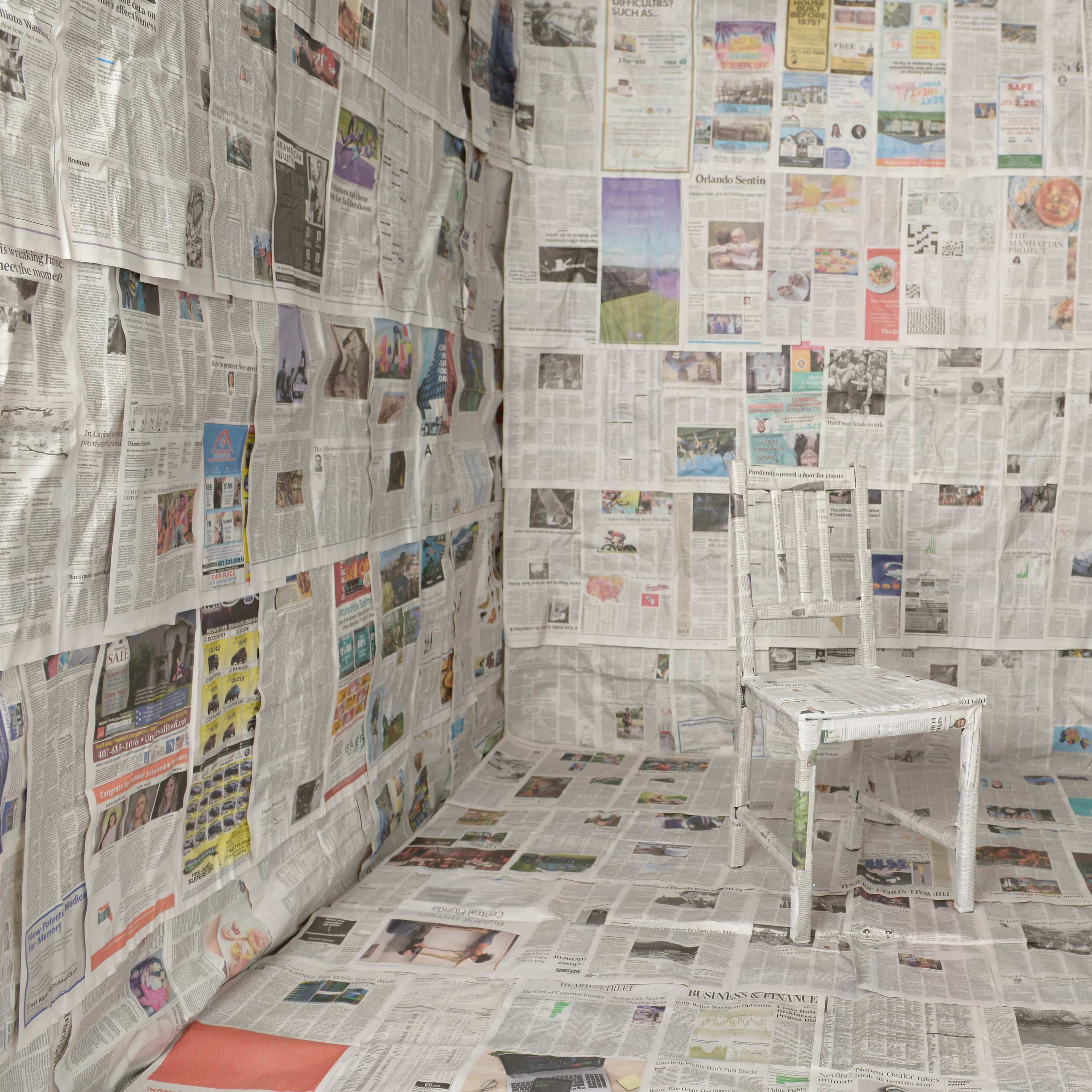 A single white chair is placed in the center of a room where the walls and floor are covered entirely in newspaper.