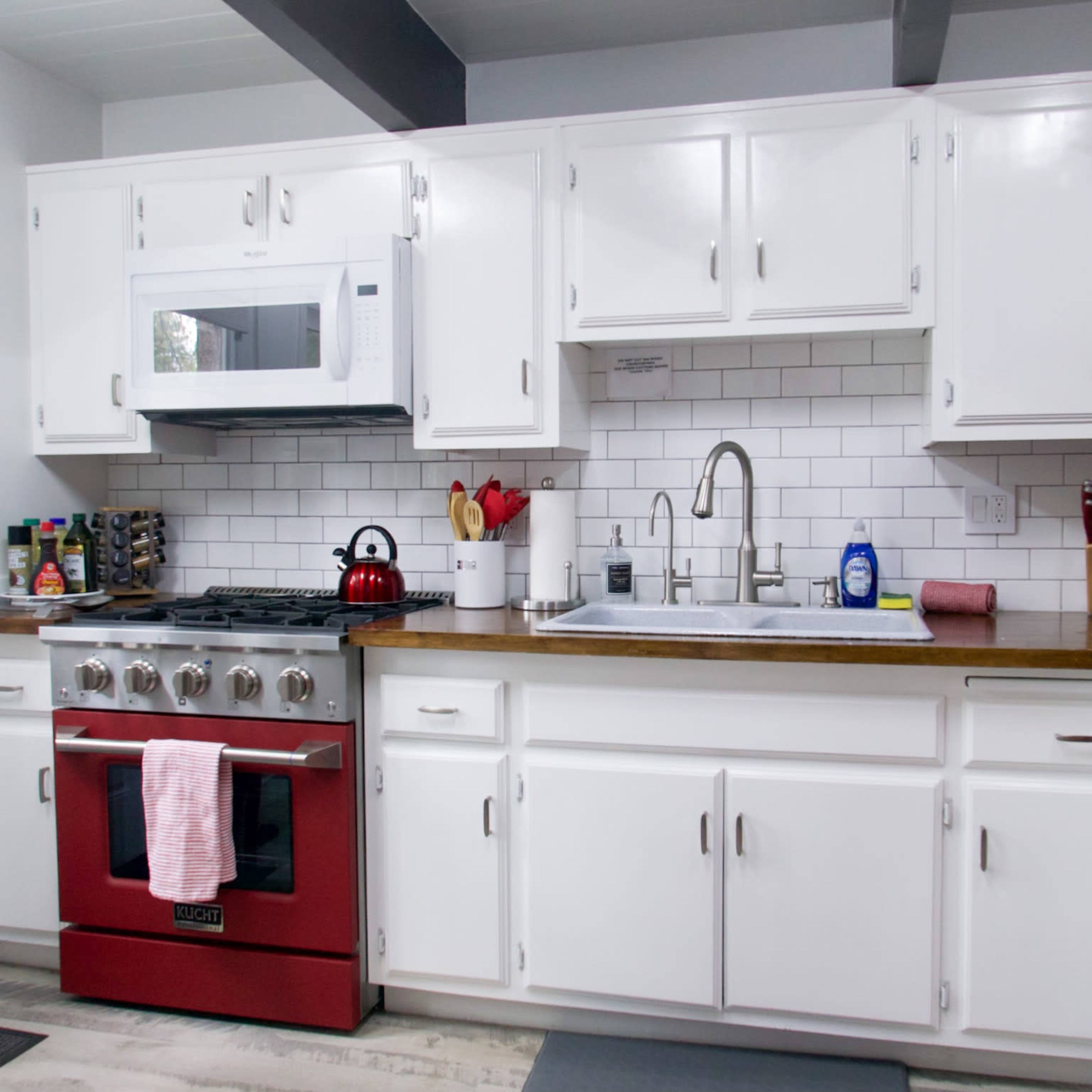 A modern kitchen featuring white cabinets, a stainless steel sink, a red stove, and a microwave above the counter, with a backsplash of white tiles.