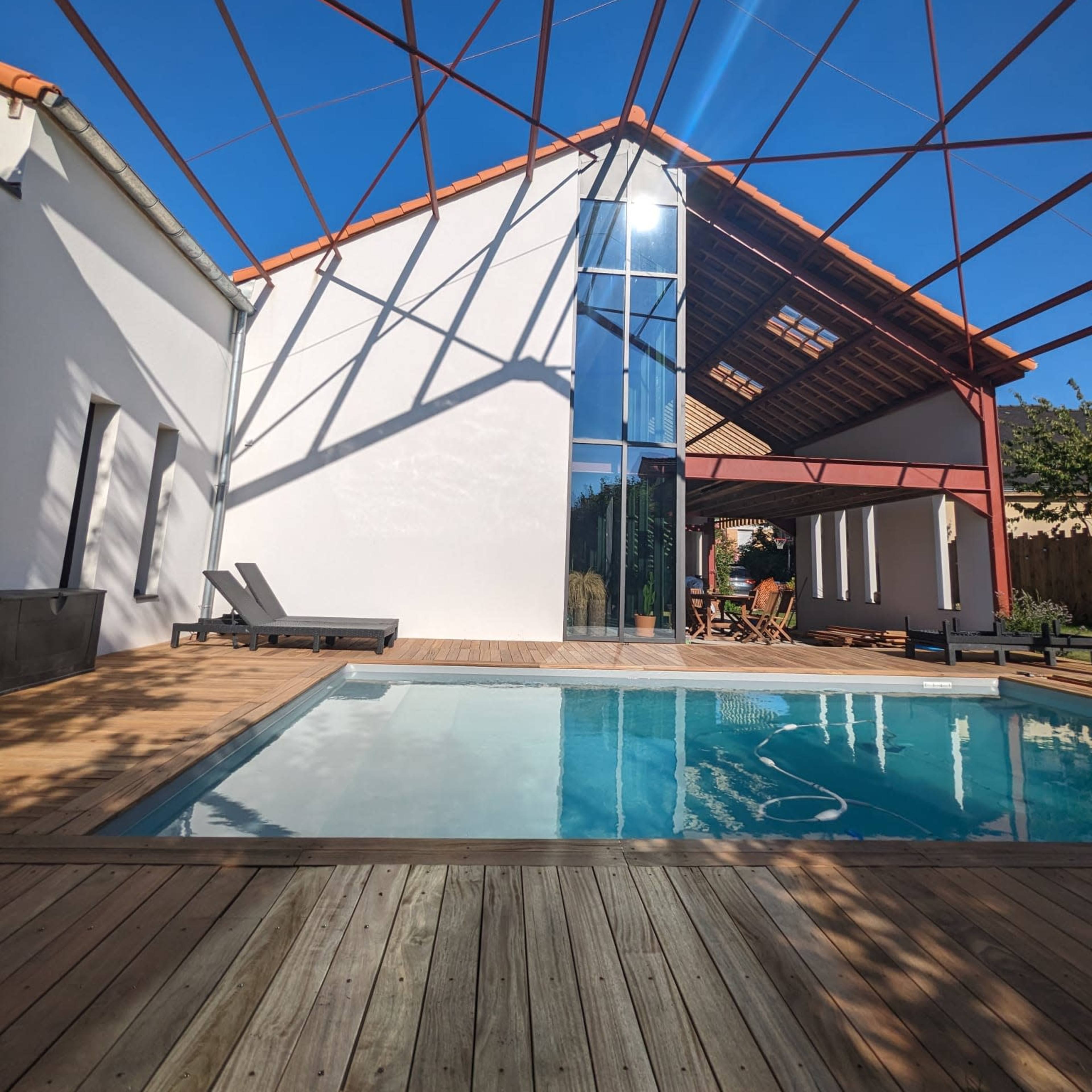 A modern building with a large glass facade, a swimming pool in the foreground, and a wooden deck under a clear blue sky with a steel frame structure overhead.