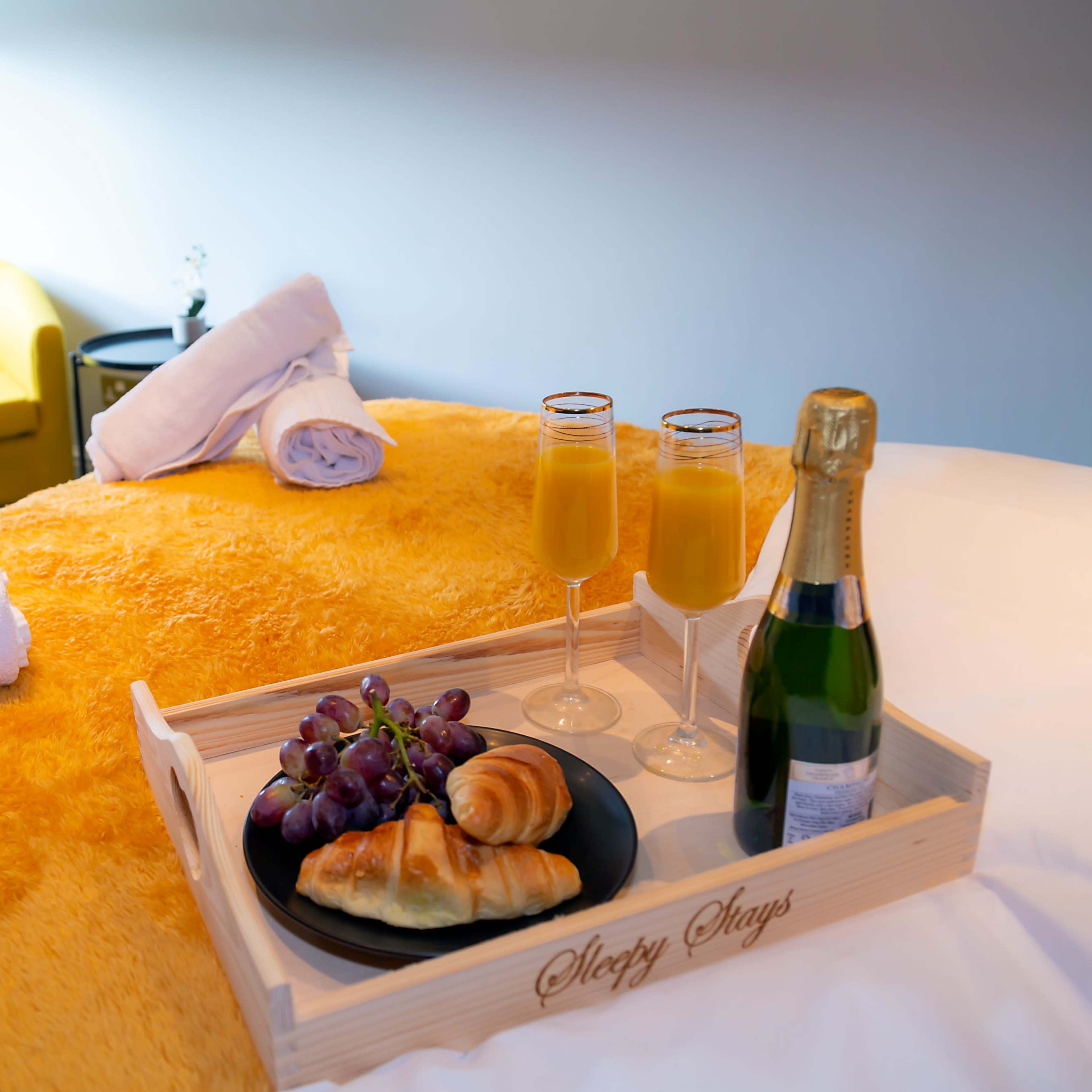 A breakfast tray with croissants, grapes, and champagne sits on a bed draped in a yellow blanket, alongside rolled towels and a chair in the background.