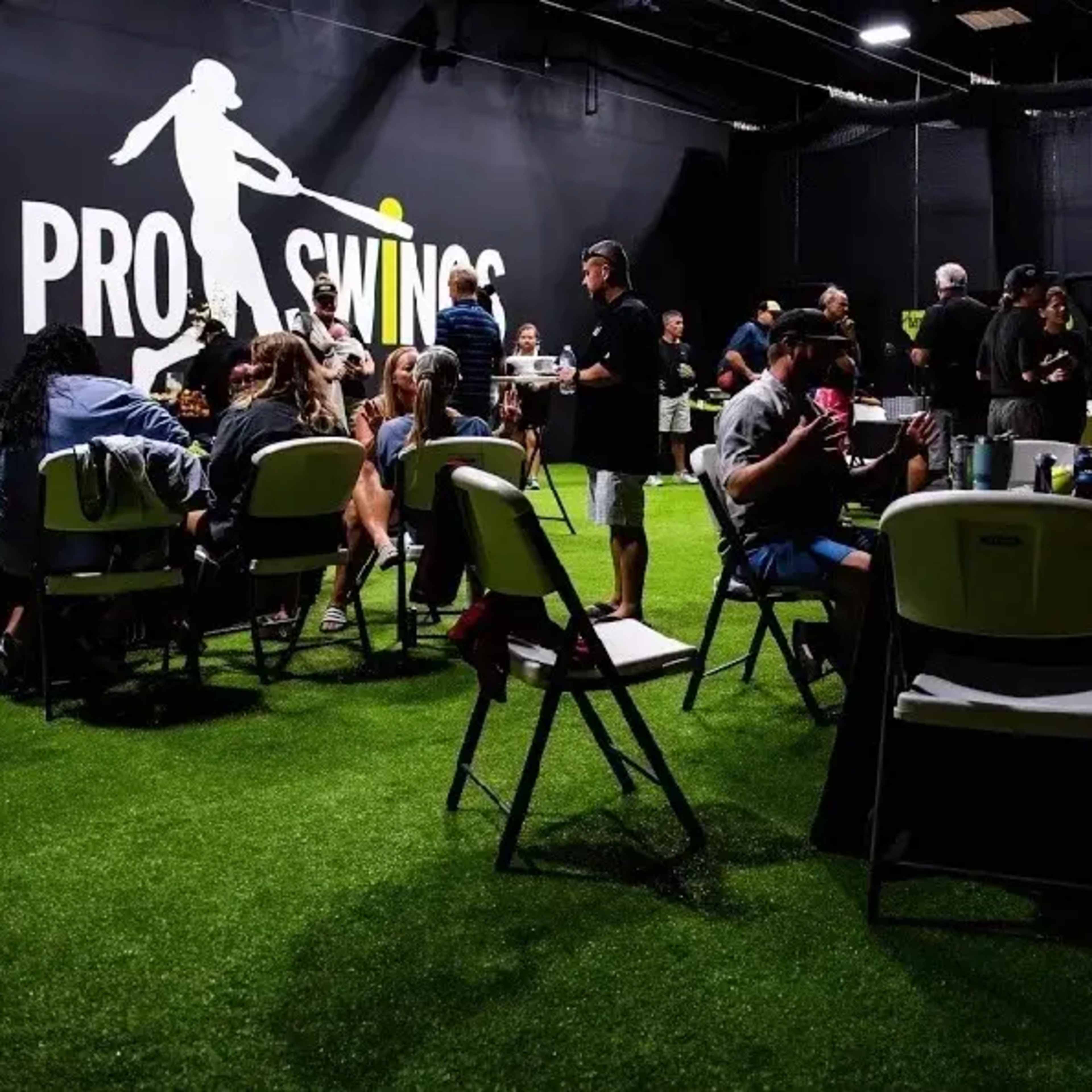 A group of people is gathered indoors at a facility called Pro Swings, engaging in various activities around tables on artificial grass.