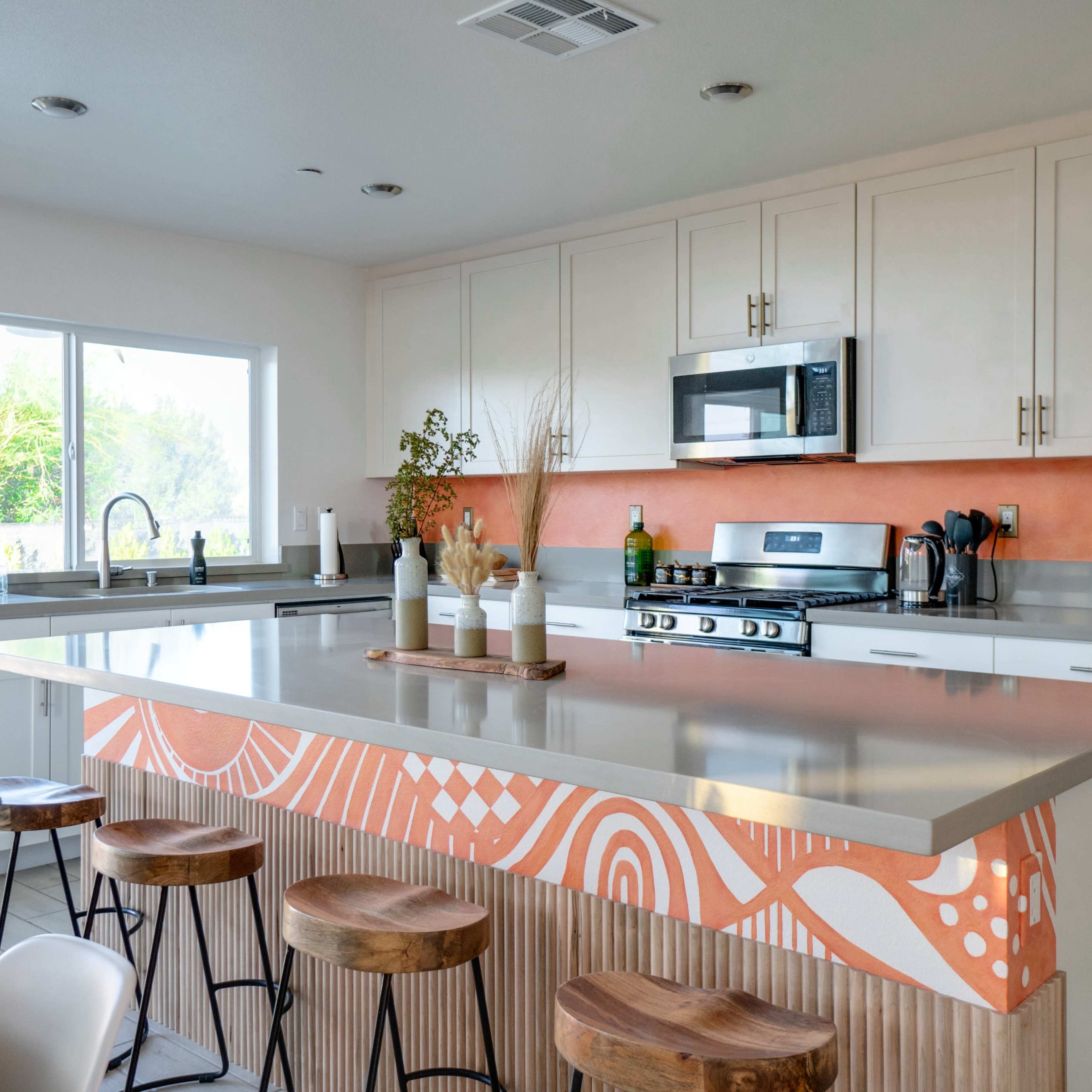 A modern kitchen features a large island with wooden stools, stainless steel appliances, and a bright orange accent wall.