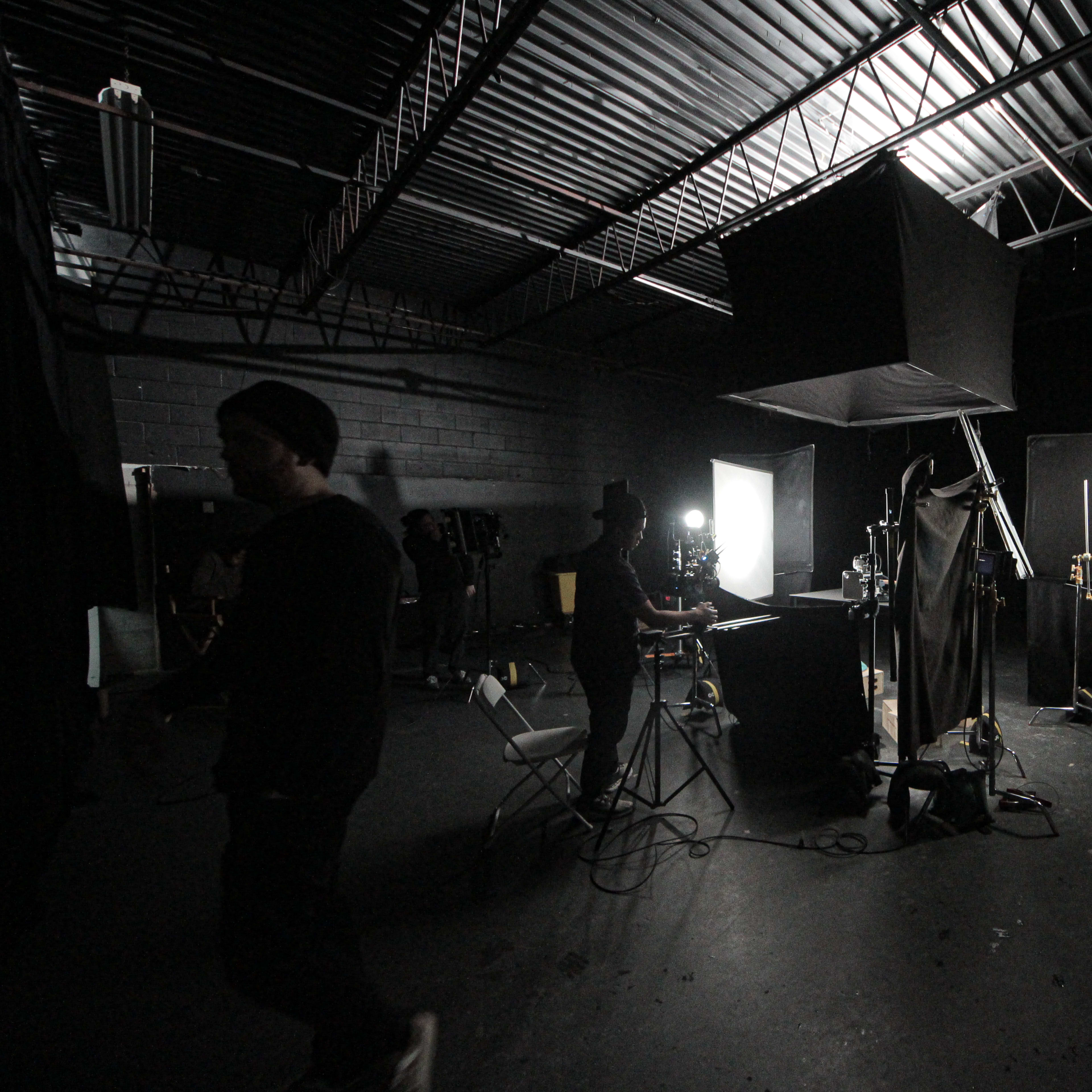 A dimly lit studio environment features various lighting setups and equipment, with individuals working amidst black curtains and backdrops.