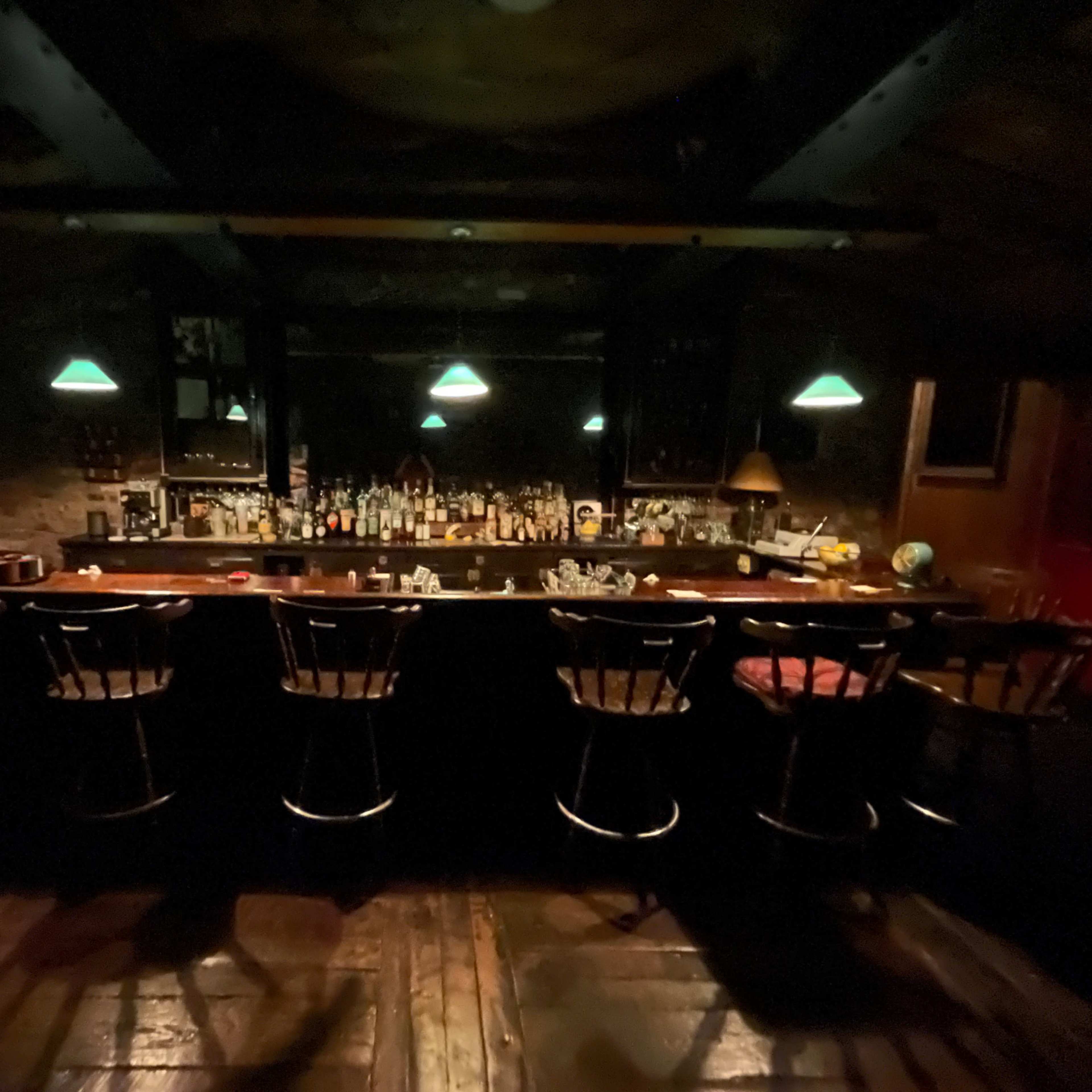 A dimly lit bar with a long wooden counter, several stools, and shelves lined with various bottles.