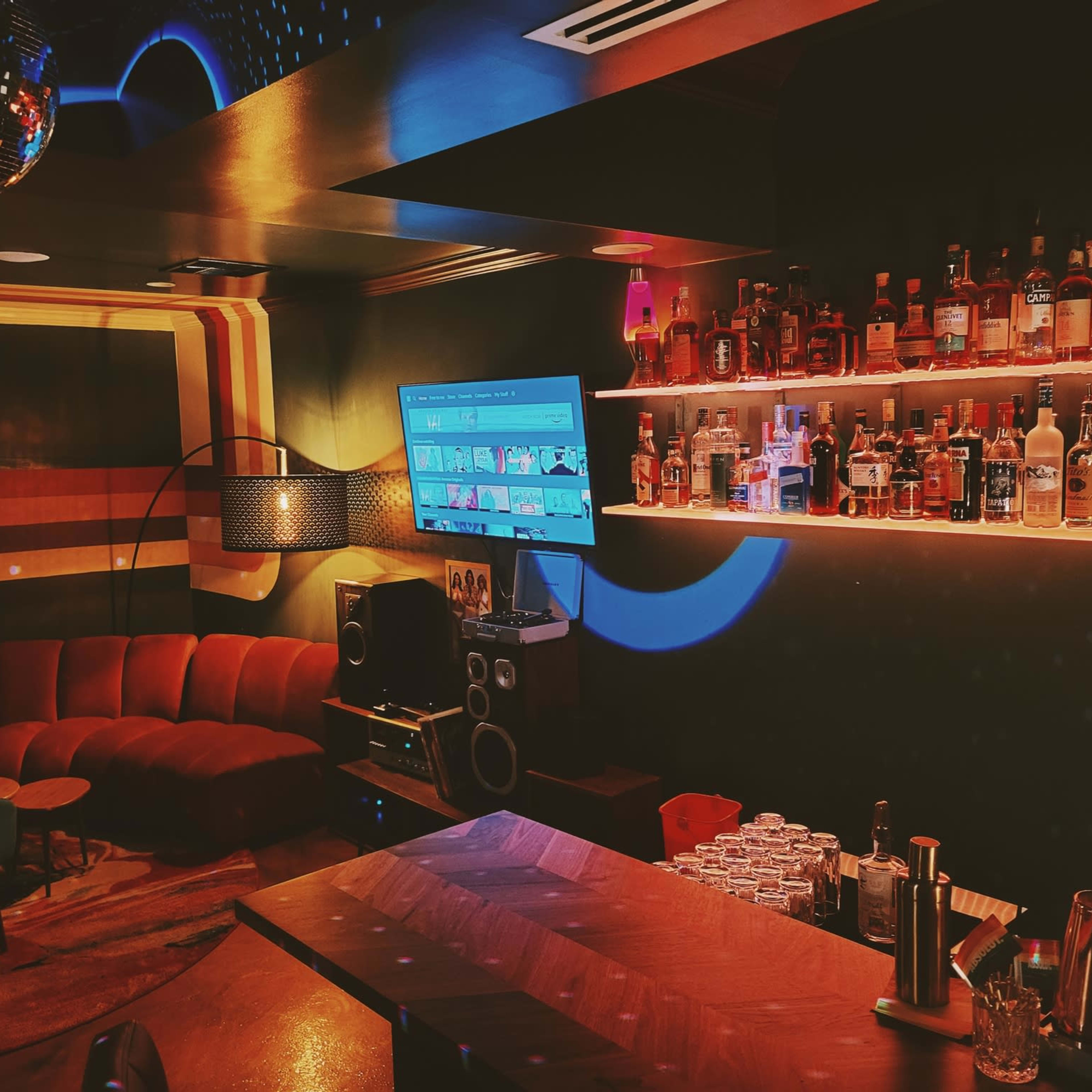 A cozy bar area features a curved red sofa, a well-stocked shelf of liquor bottles, and a large television on the wall.