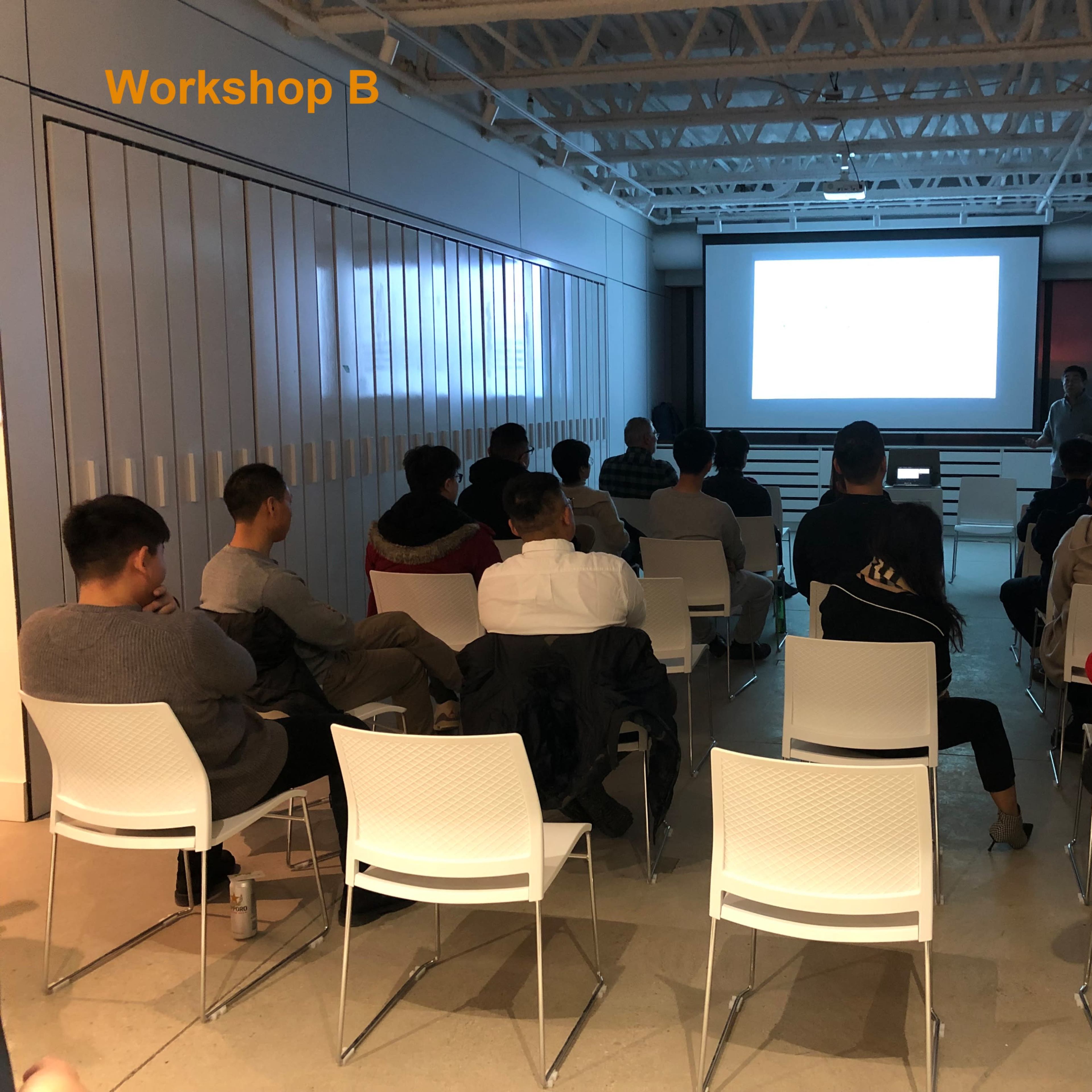 A group of people sits in white chairs facing a presentation screen in a workshop environment.