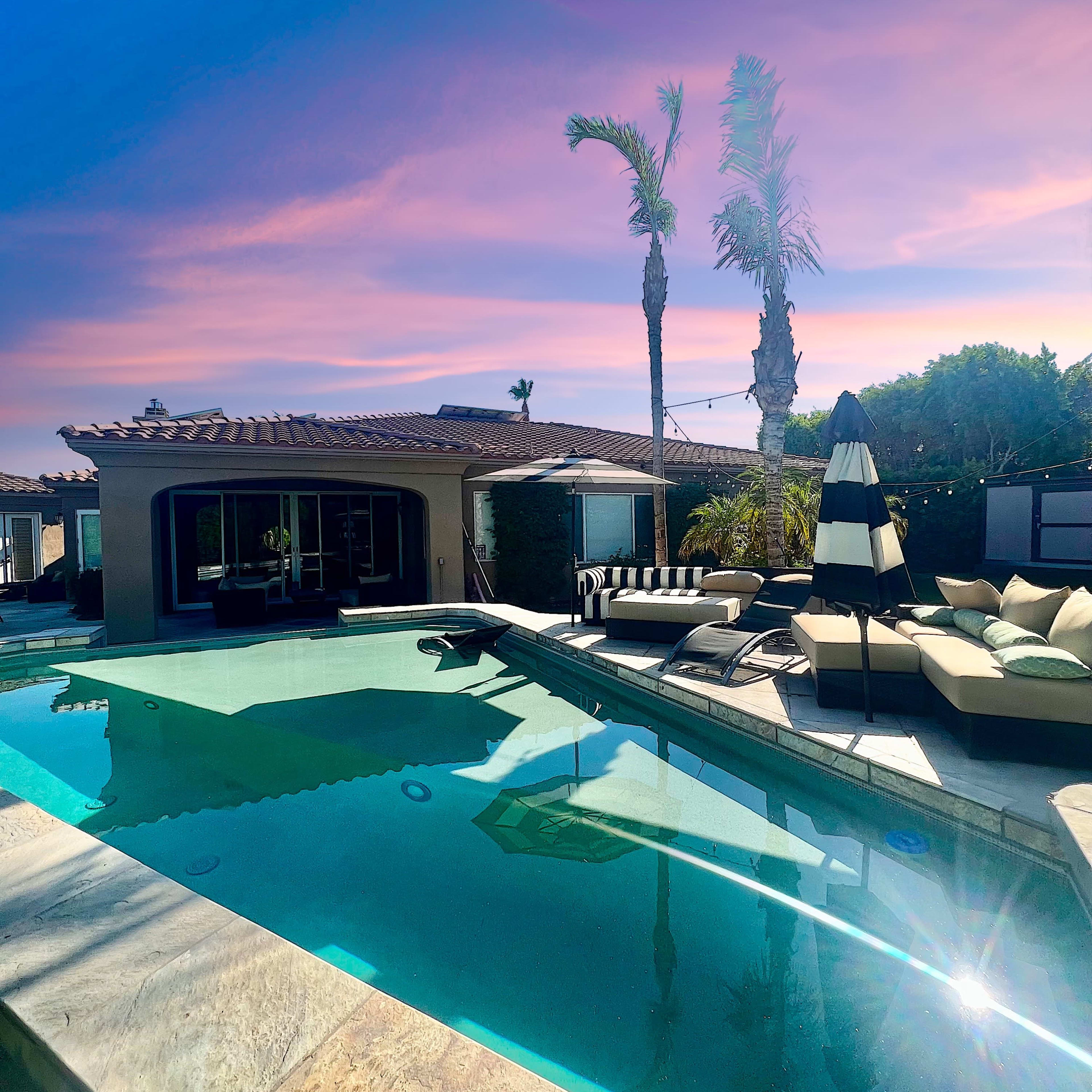 A swimming pool surrounded by lounge chairs and umbrellas, with palm trees and a home in the background under a colorful sky.