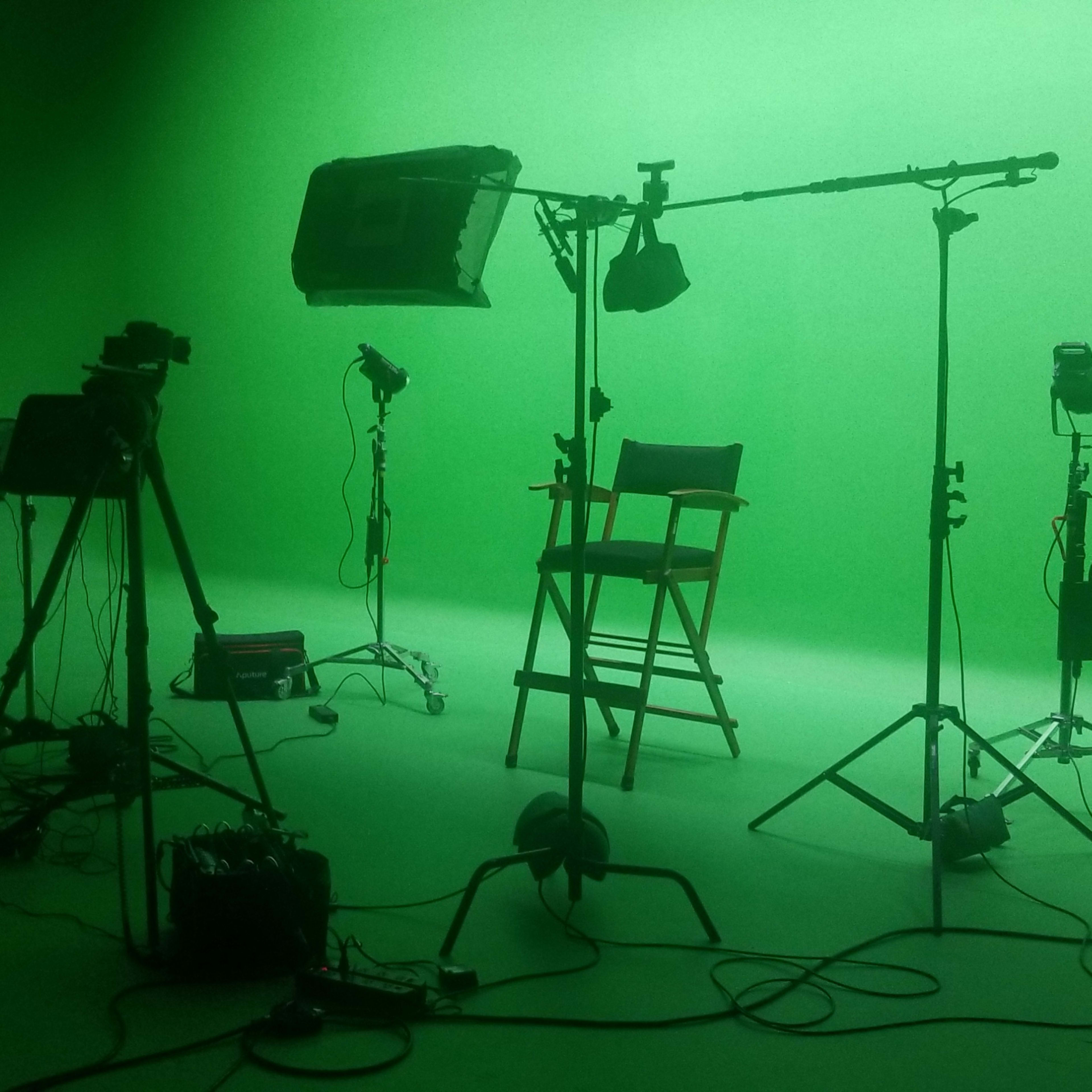 A film studio is set up with several lights, cameras, and a green backdrop, along with director's chairs arranged for a shoot.