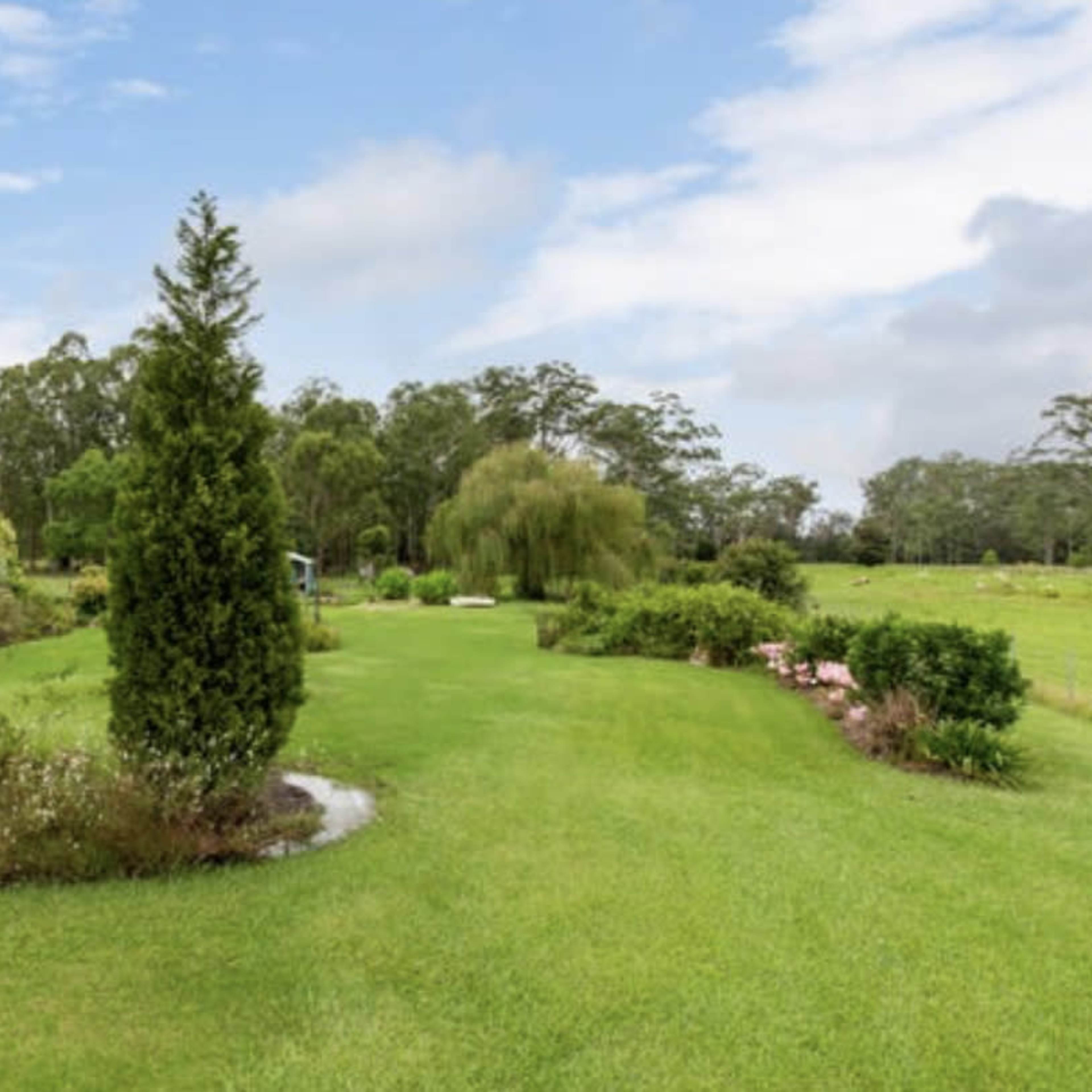 A lush garden with neatly trimmed grass, various plants, and trees under a partly cloudy sky.