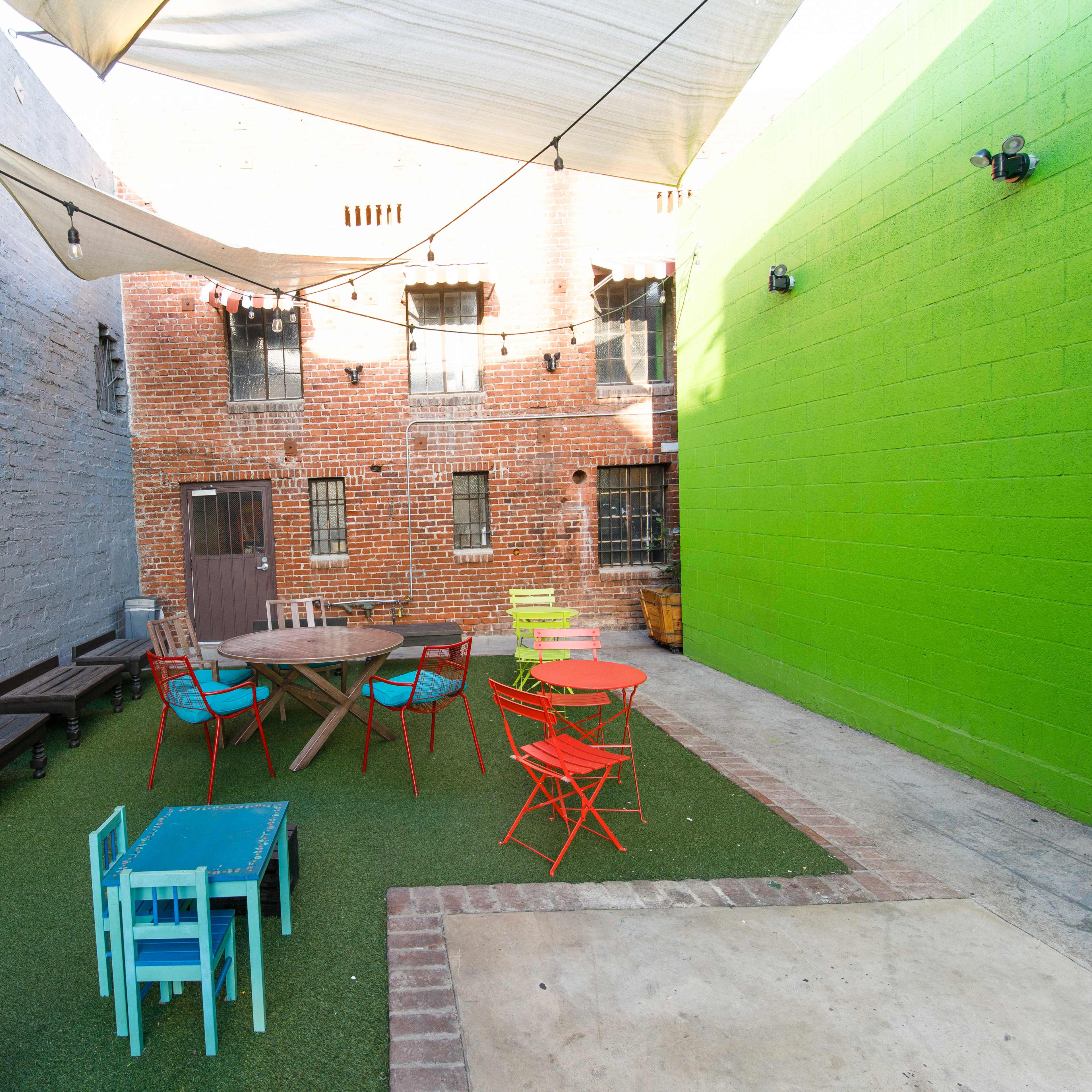 The scene features a small outdoor courtyard with colorful furniture, including a round table surrounded by chairs, bordered by gray brick walls and a vibrant green wall.