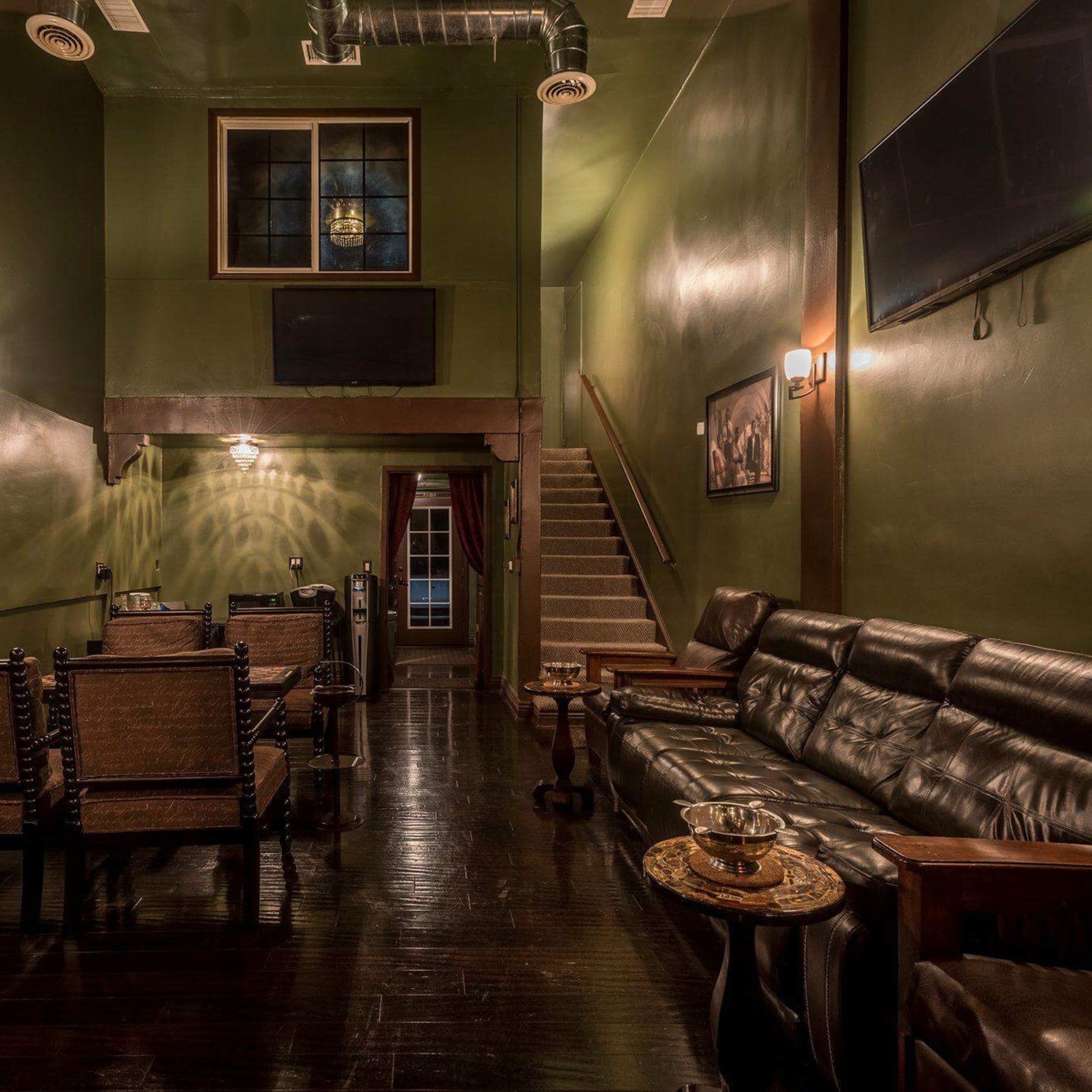 A dimly lit lounge with green walls, black wooden floors, leather seating, and a staircase leading to a second level.
