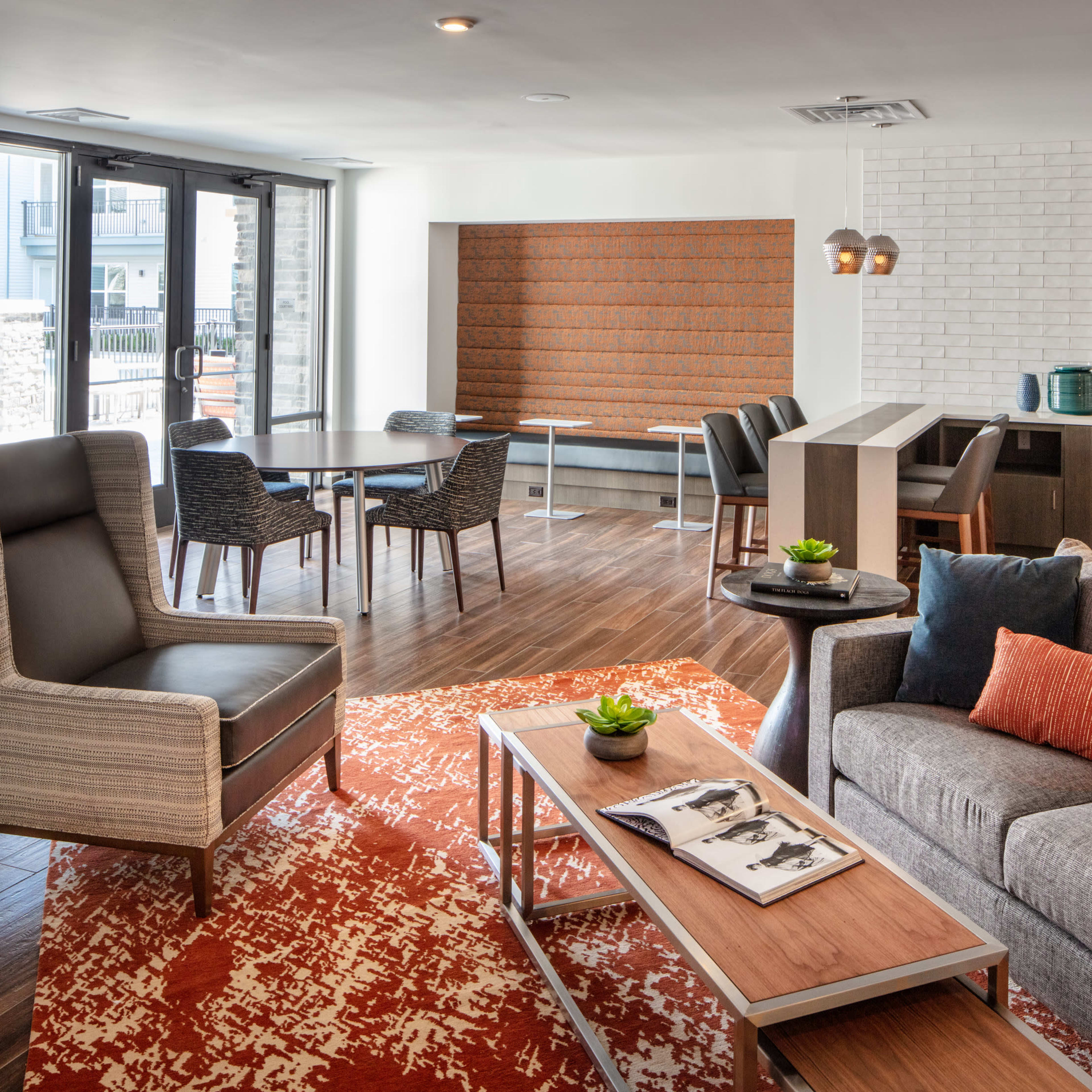 A modern living area with a gray sofa, orange accent pillows, a coffee table, and a dining space featuring a round table and chairs, all set in a well-lit, open-layout room with large windows.