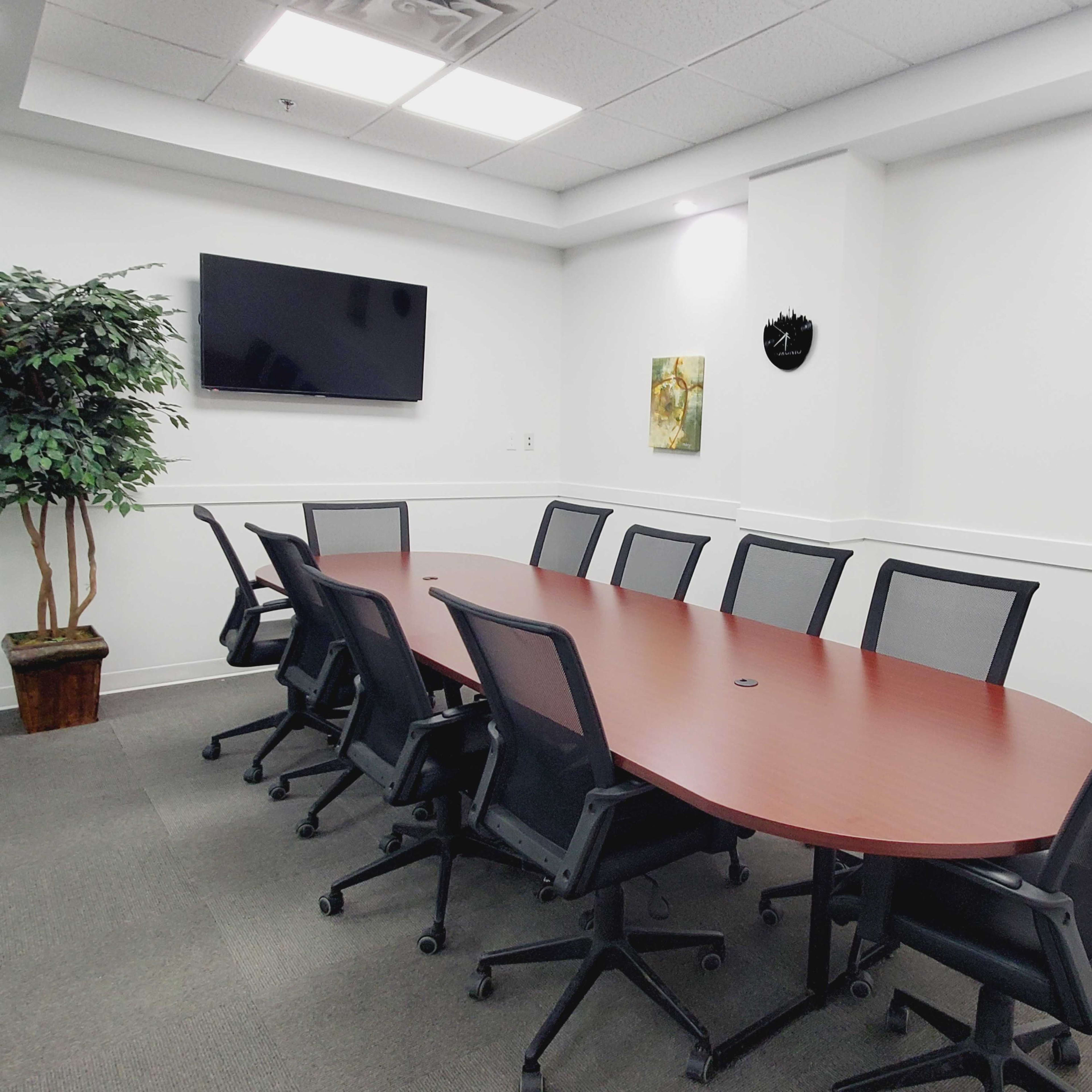 A conference room features an oval wooden table surrounded by black chairs, with a TV and two framed artworks on the walls.