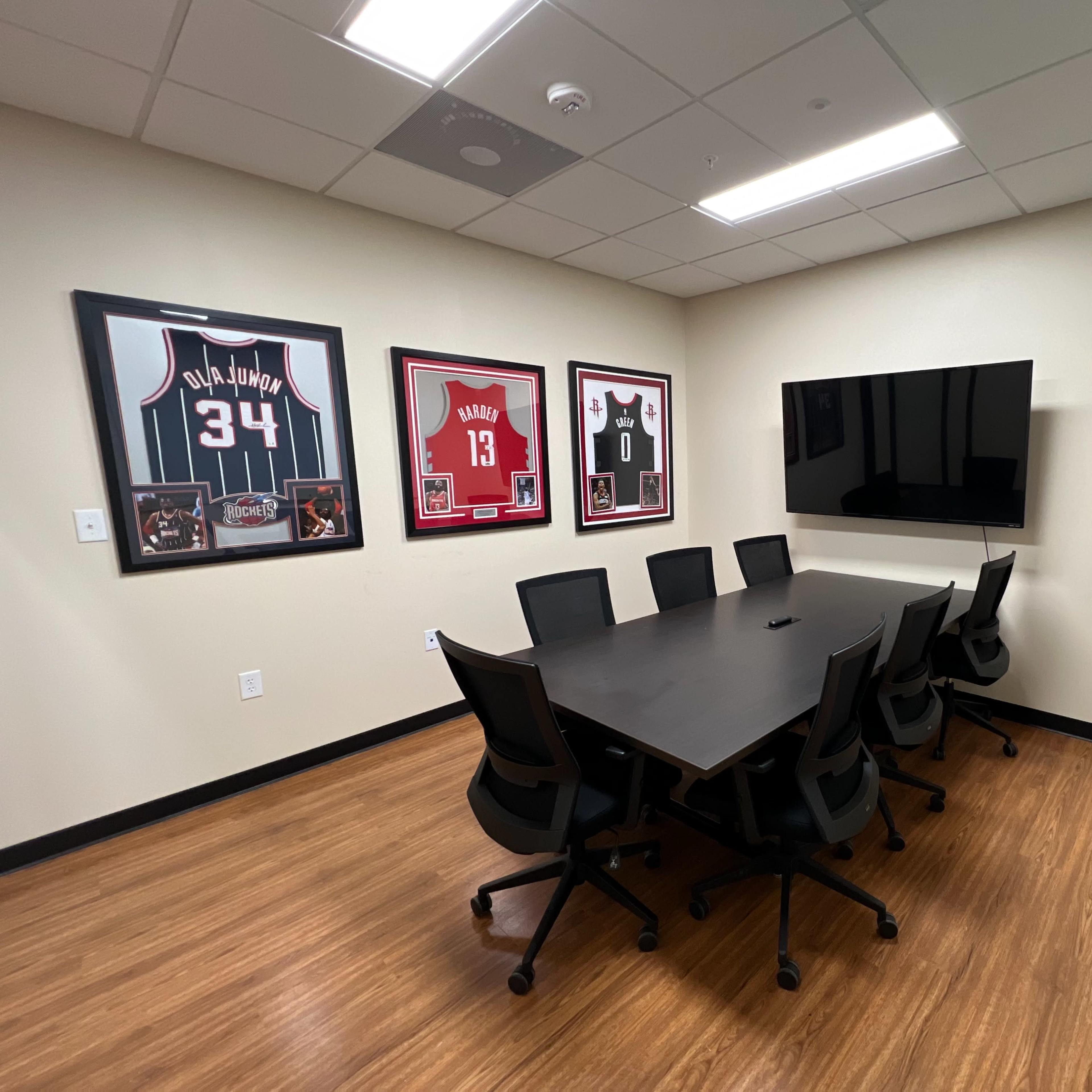 A conference room features a long black table surrounded by chairs, with sports jerseys displayed on the walls and a wall-mounted television.