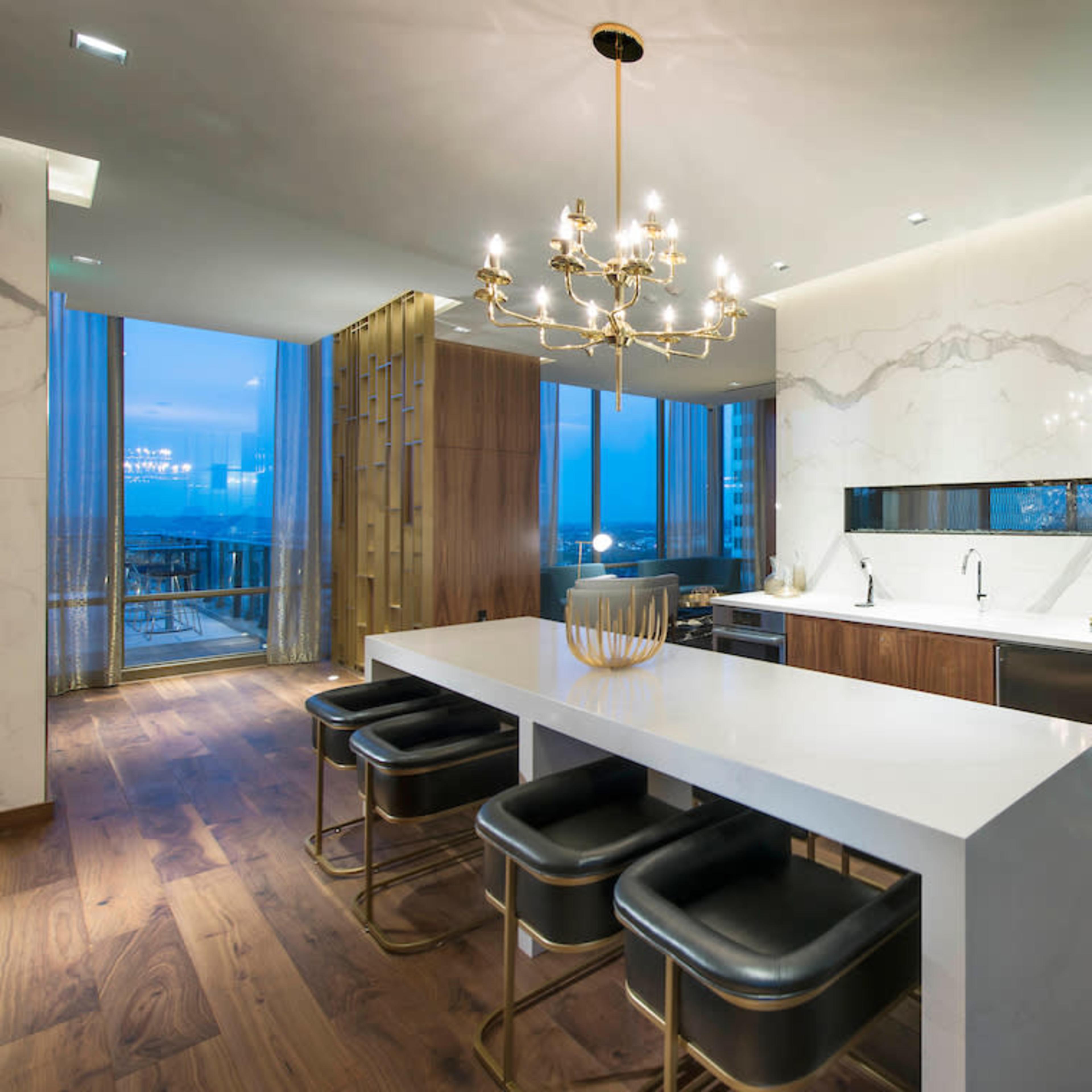 A modern kitchen features a large island with black stools, elegant light fixtures, and large windows offering a view of the city.