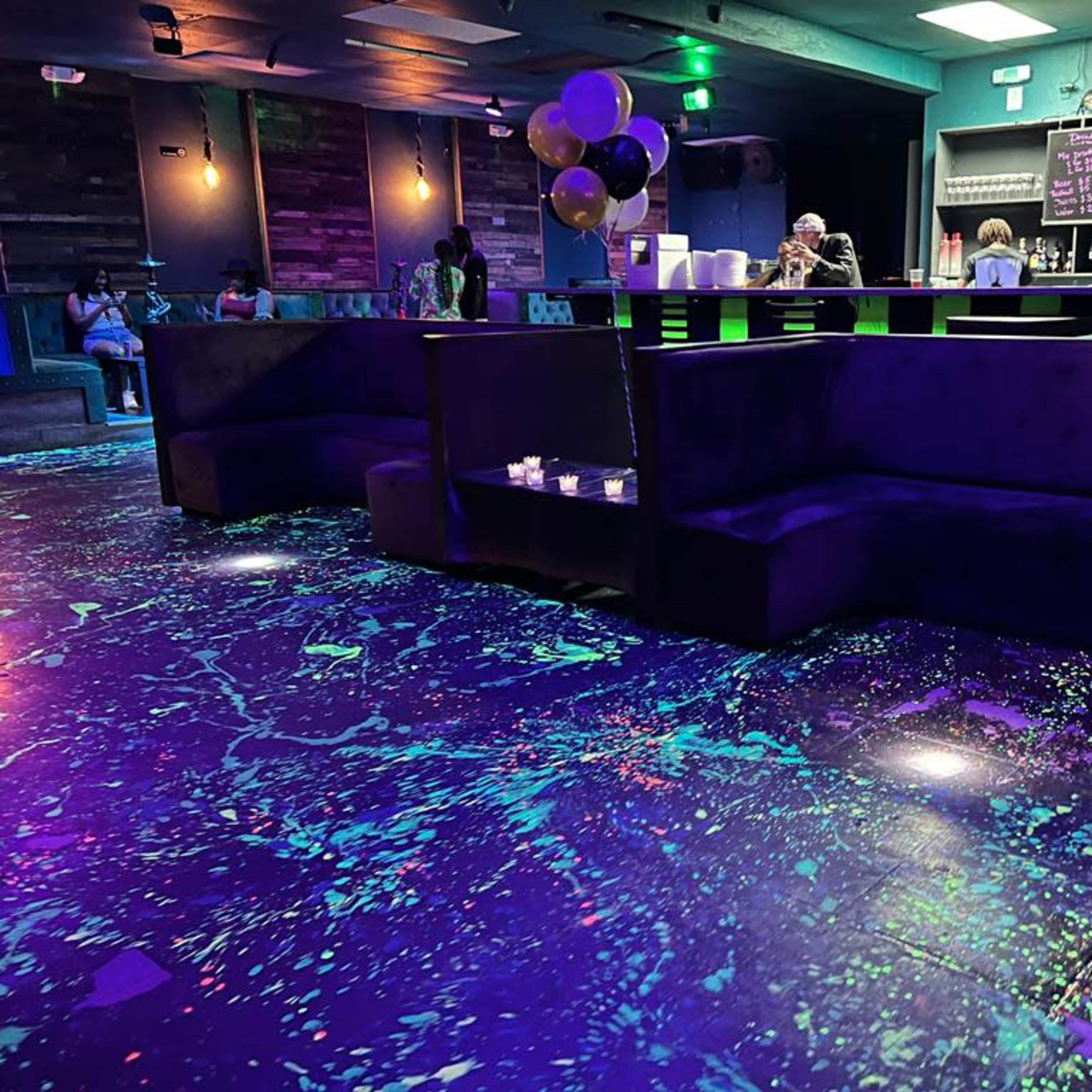 A dimly lit lounge with dark seating, a bar area, and a colorful, splattered floor.
