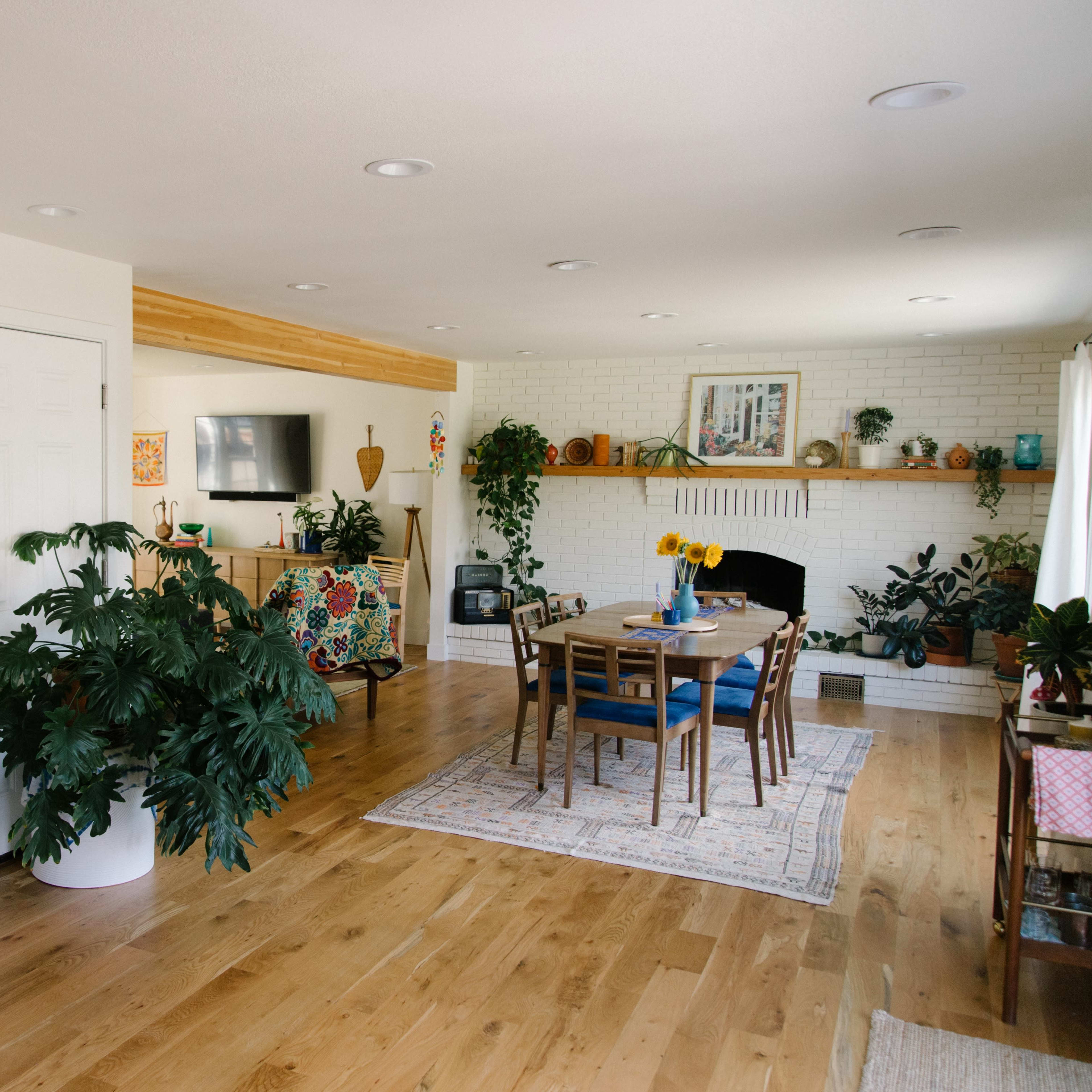 A bright living space features a dining table centered under overhead lights, surrounded by plants, a television, and a cozy fireplace with a light-colored brick surround.