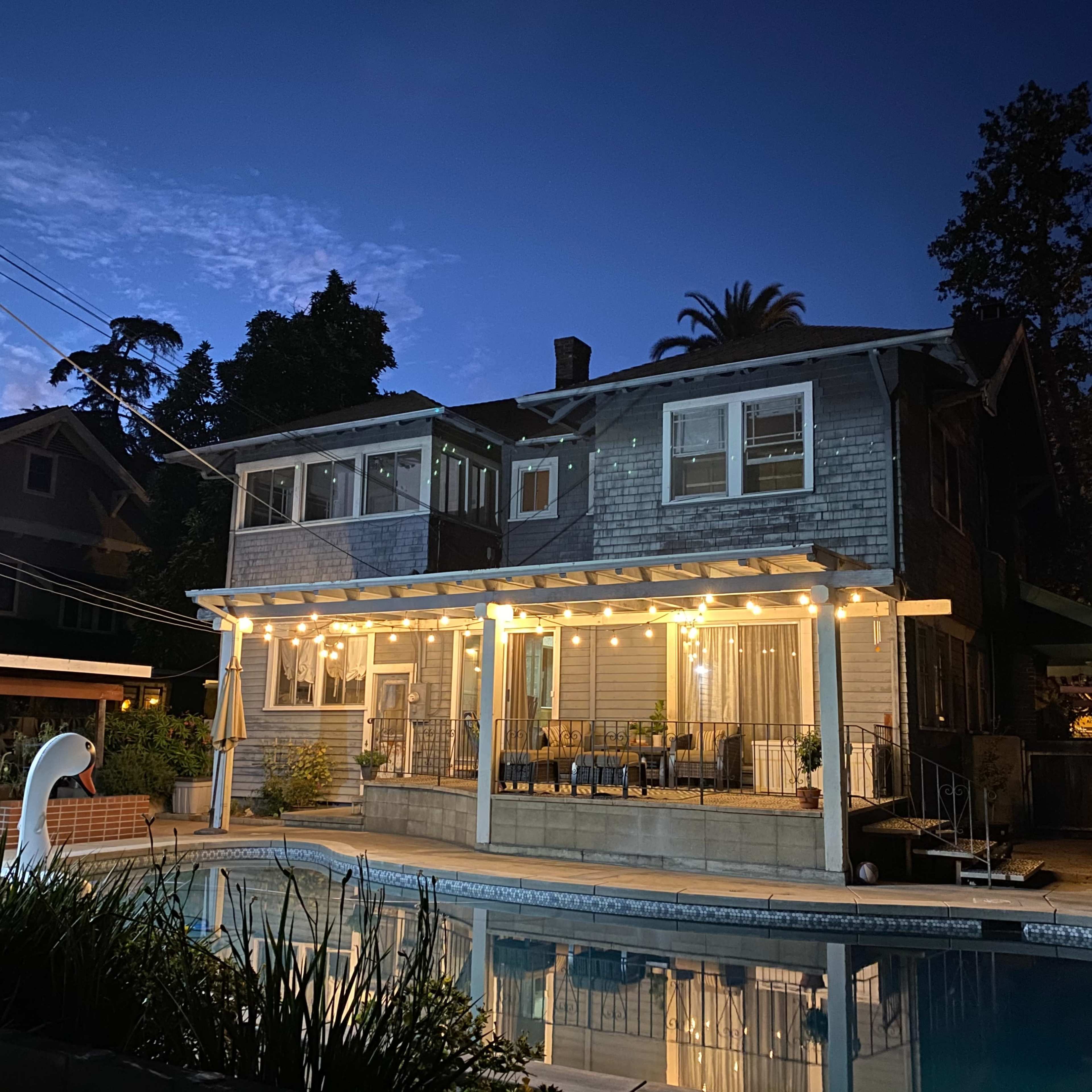 A two-story house with outdoor lighting and a swimming pool is set against a twilight sky.
