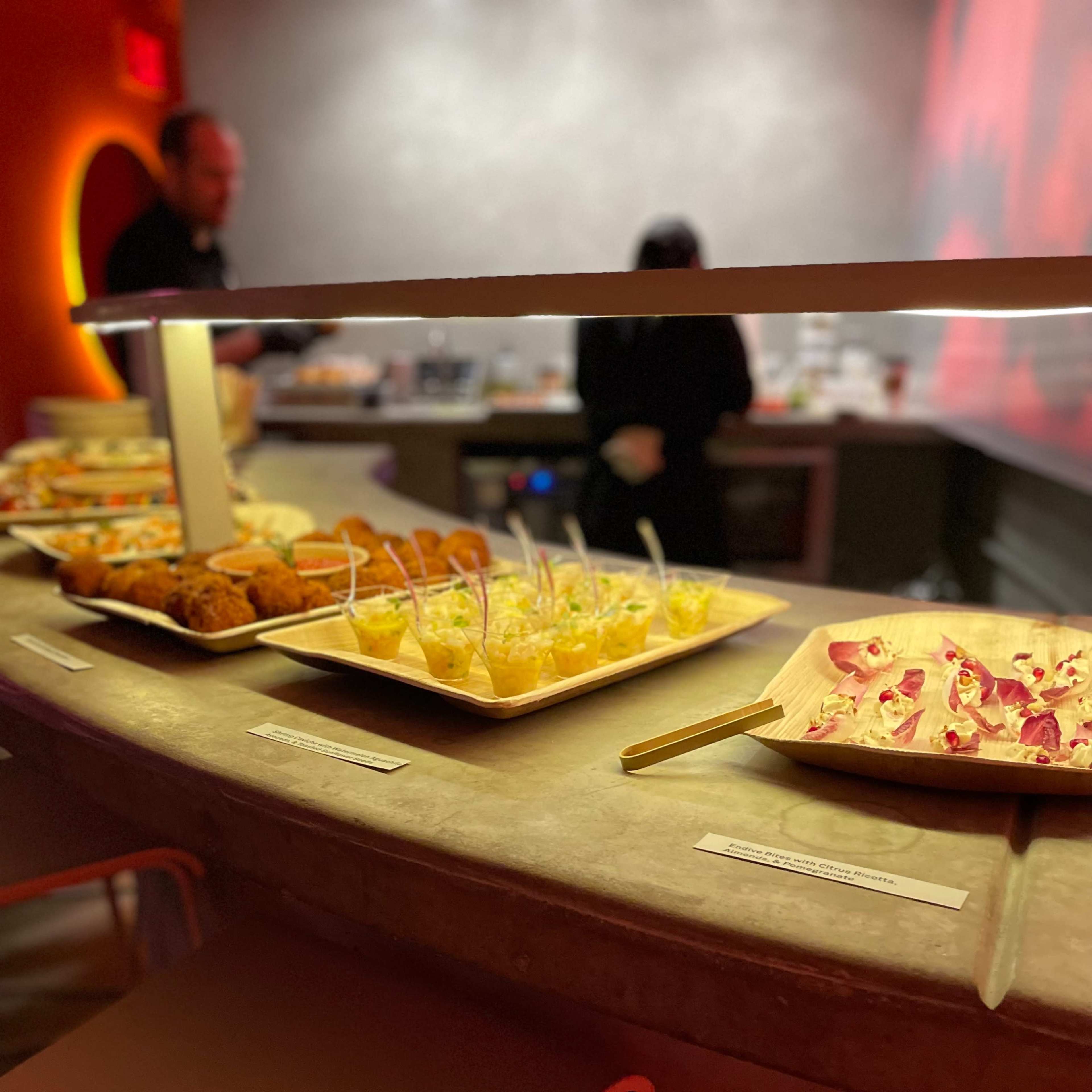 A buffet table displays various appetizers and small dishes, with a modern bar area in the background.