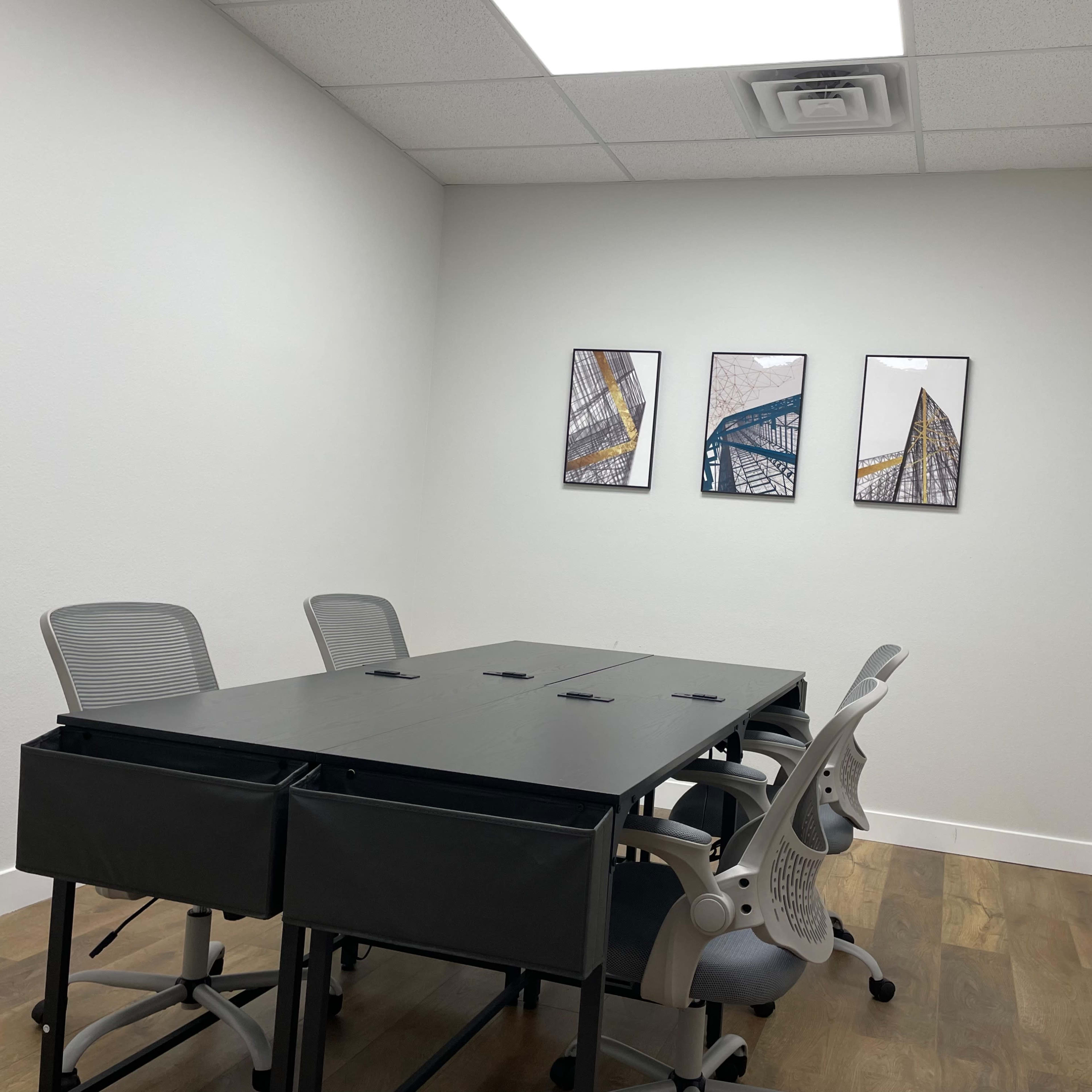 A small conference room with a rectangular table and four ergonomic chairs, featuring three framed artworks on the wall.