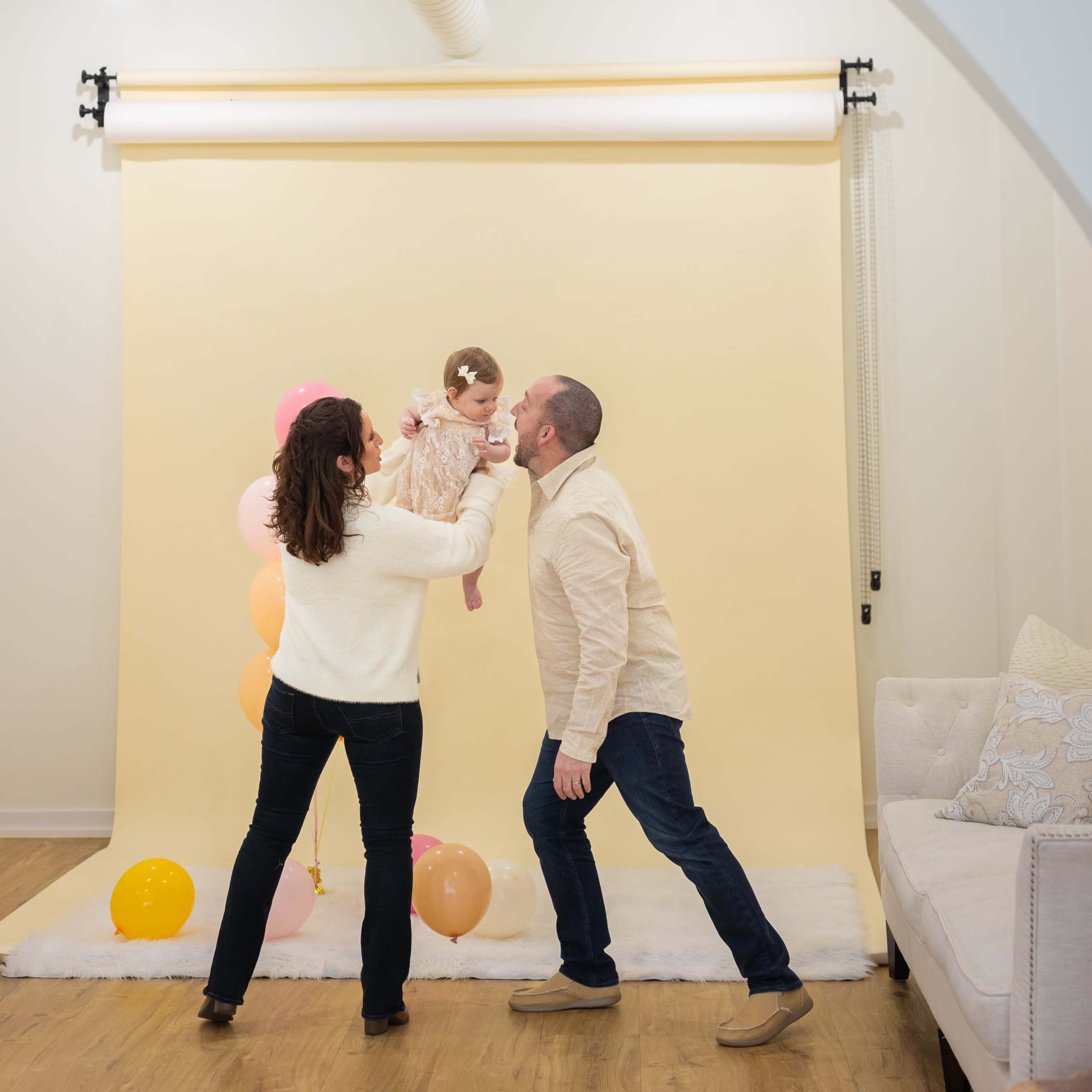 A couple lifts their child in front of a light backdrop decorated with balloons.